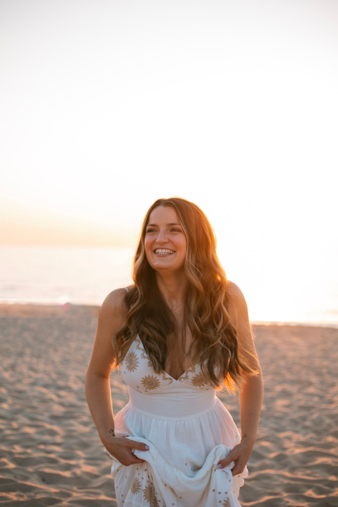 Joyful woman in white floral sundress laughing on sandy beach during golden sunset.