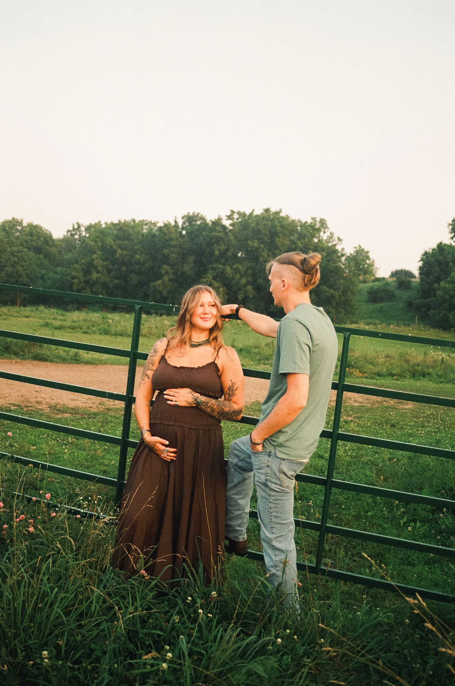 Pregnant woman in brown dress with partner touching her hair by a farm gate at golden hour.