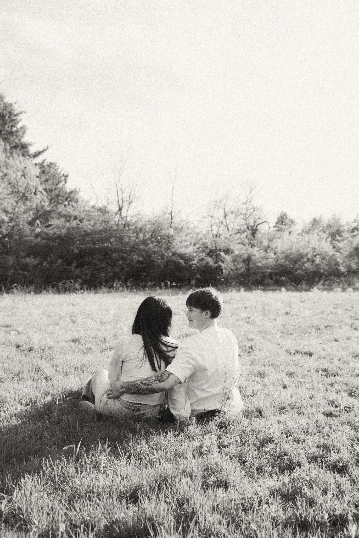 Black and white photo of couple sitting close together in a grassy field surrounded by trees.