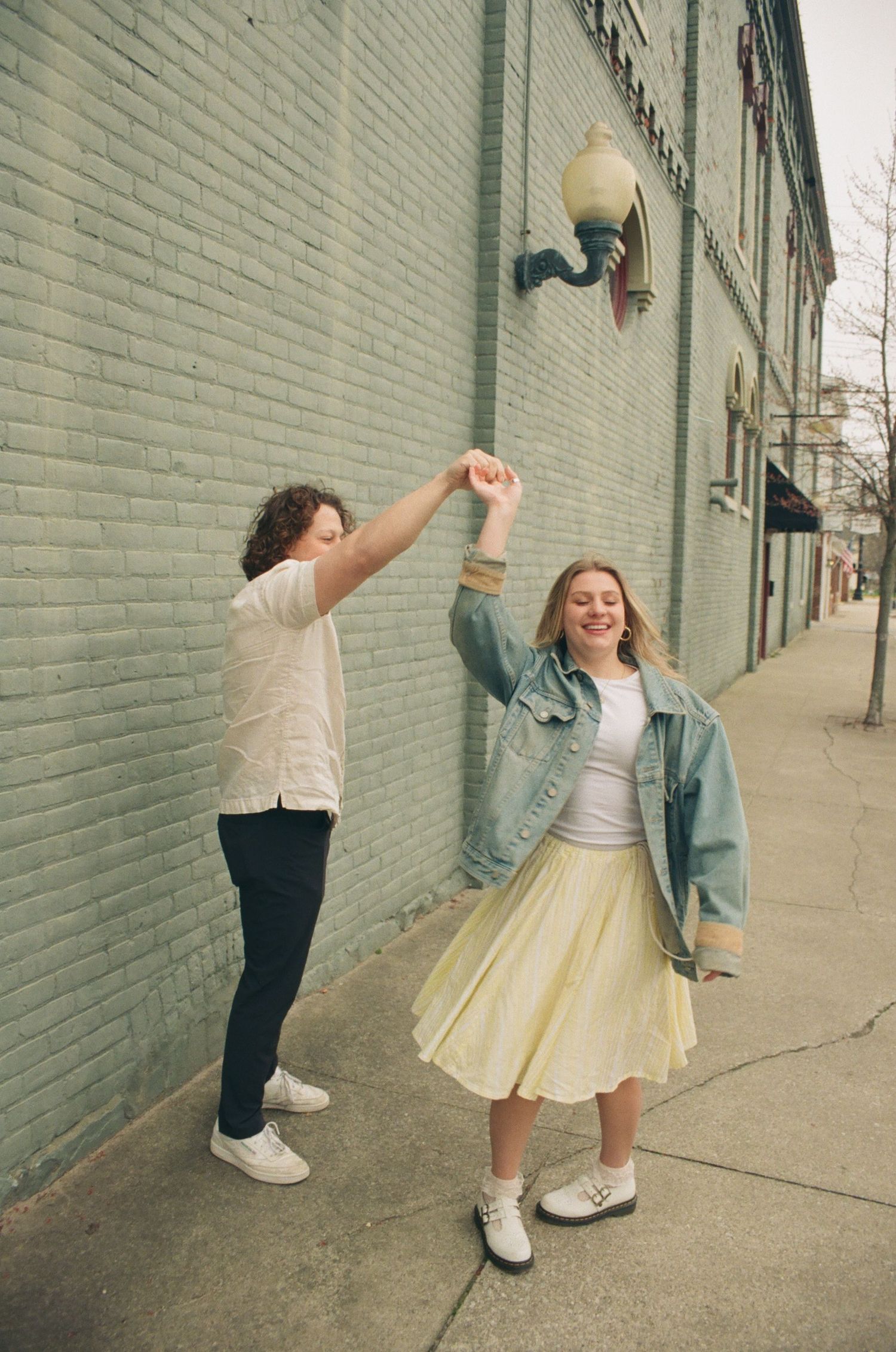 Joyful couple dancing on a sidewalk, man spinning smiling woman in yellow skirt and denim jacket.