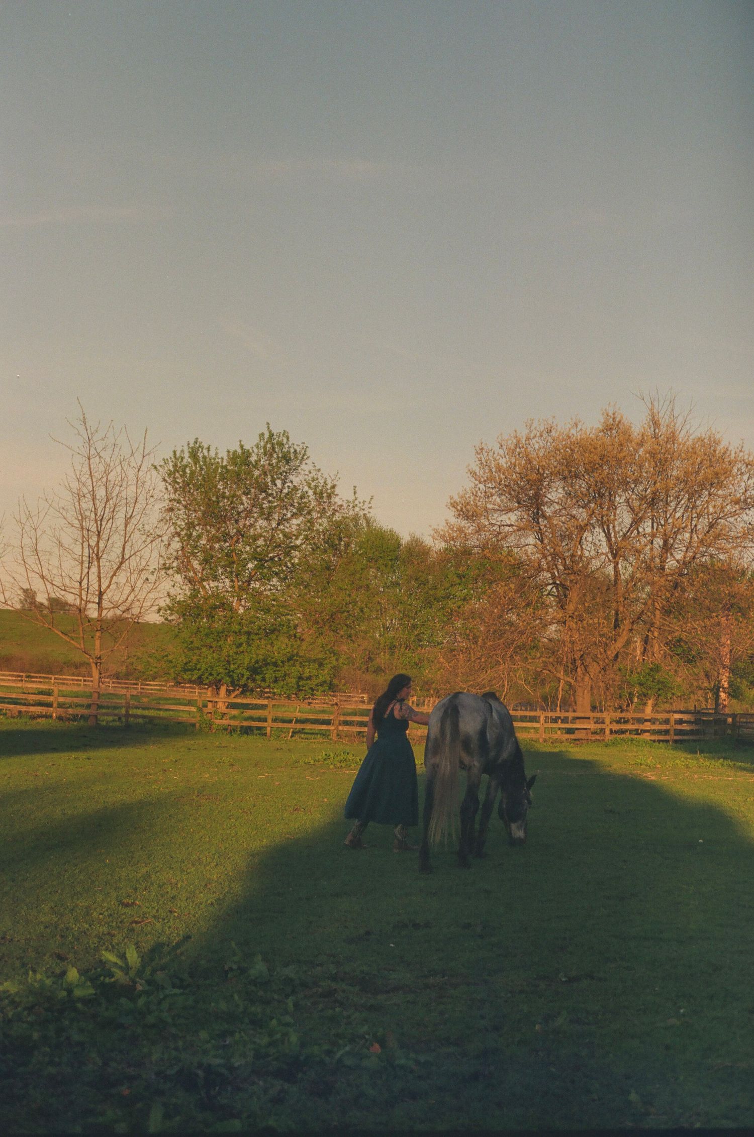 Woman in dark dress standing beside a horse grazing in a green field at golden hour.