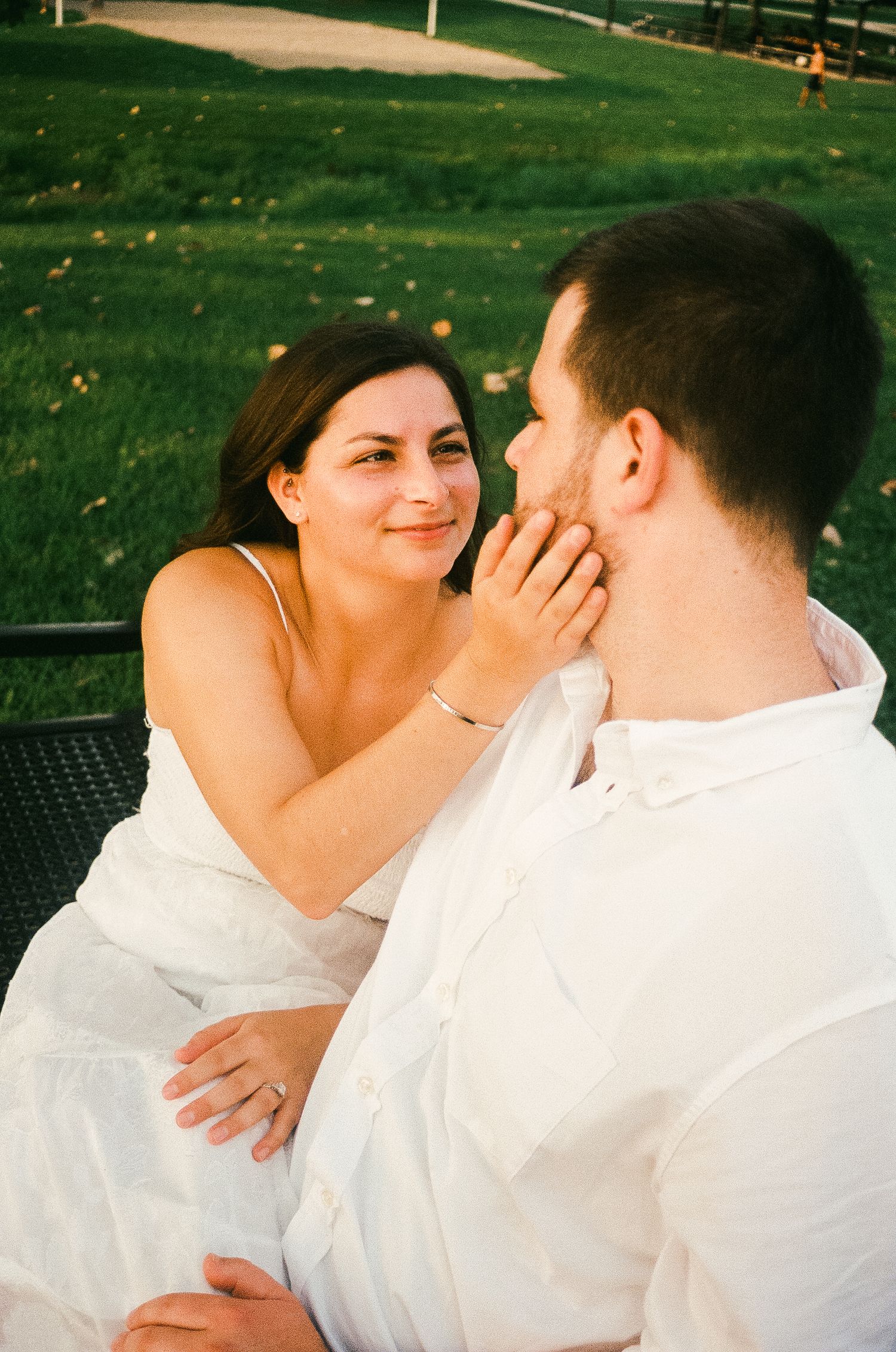 A couple in white outfits share a tender moment outdoors, with the woman gently touching the man's face while smiling.