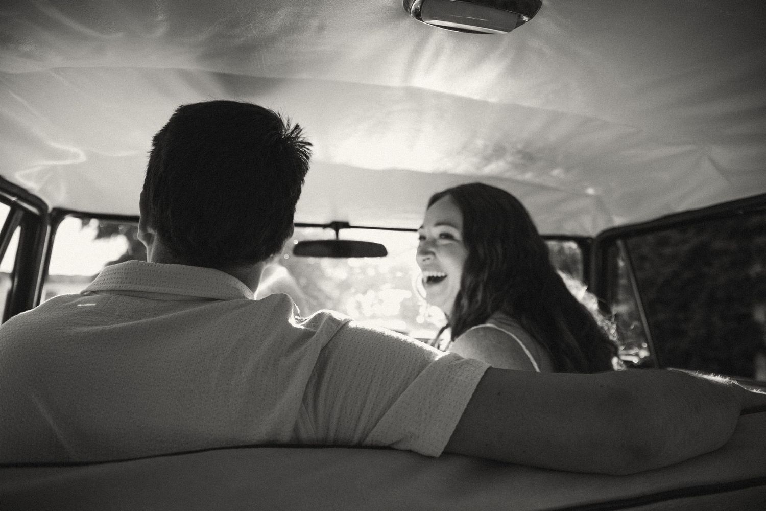 Black and white photo of a smiling couple sitting inside a vintage van, sharing a joyful moment together.