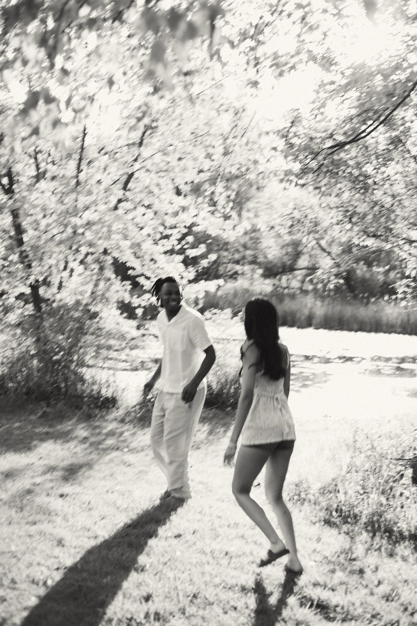 A black and white photo of a couple walking toward each other near a sunlit riverside.