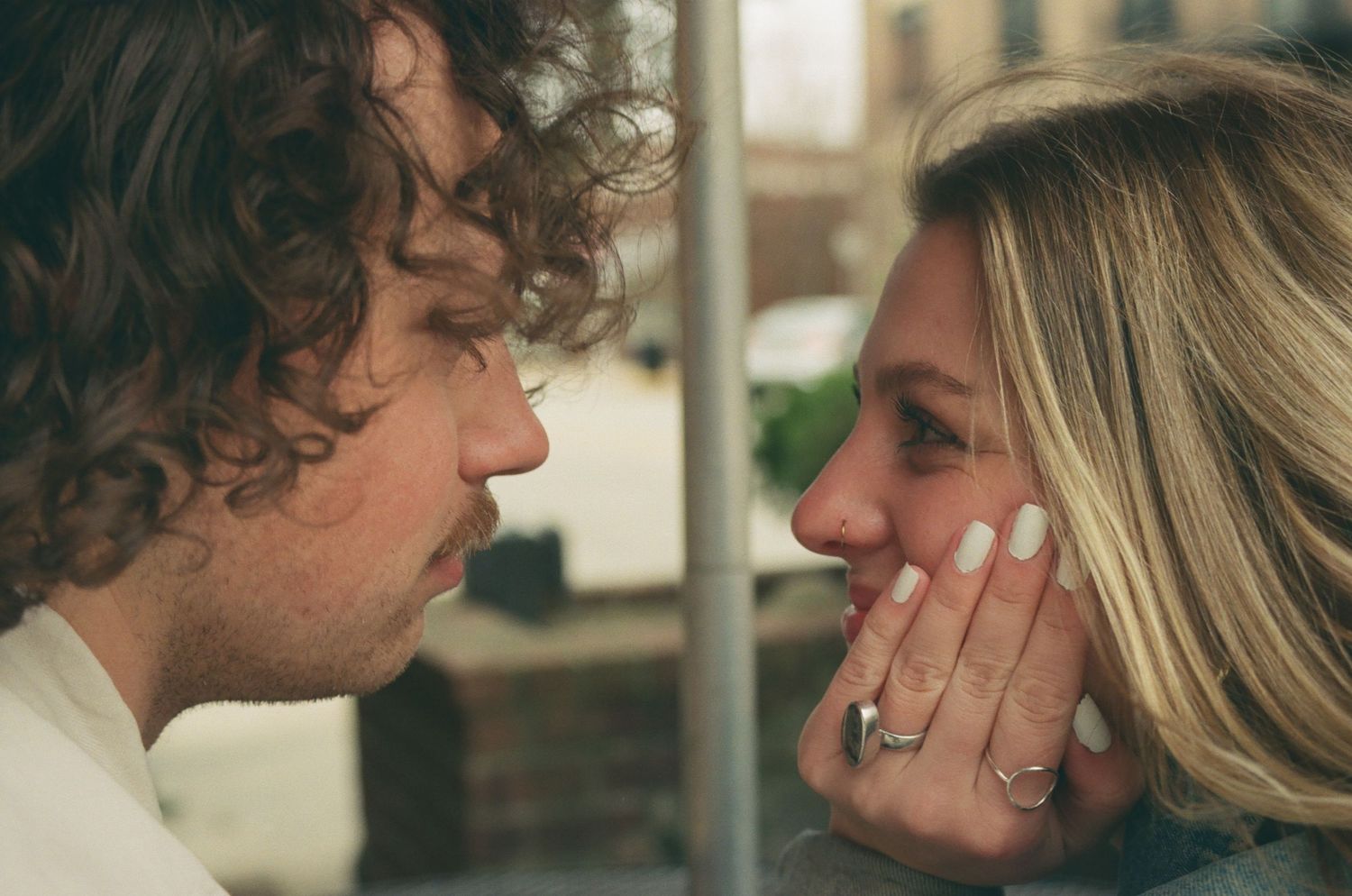 A curly-haired man and a smiling blonde woman share an intimate close-up moment face to face.