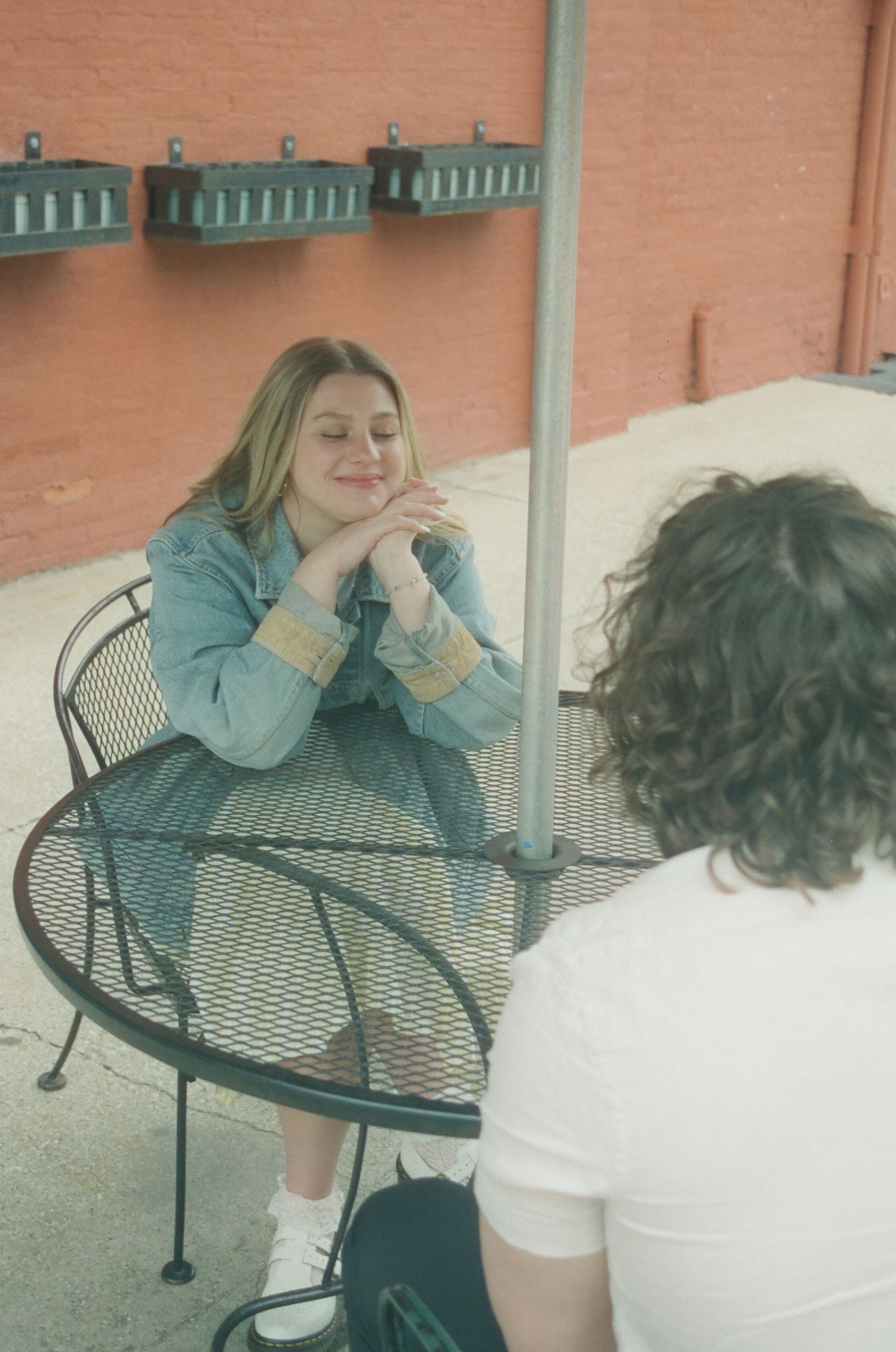Happy blonde woman in denim jacket smiling warmly at outdoor cafe table with hands folded.