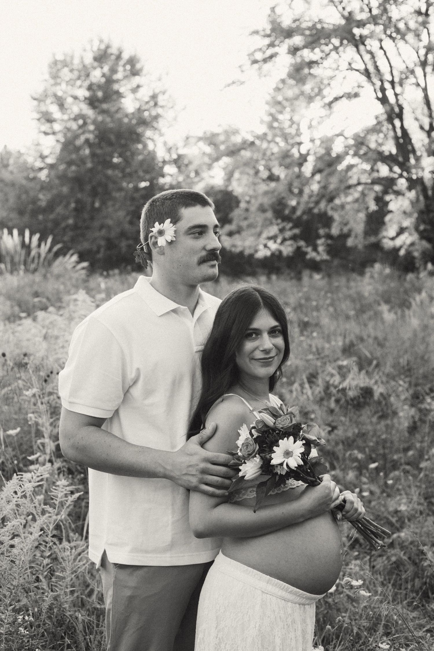 Black and white maternity portrait of couple embracing in field, woman holding wildflower bouquet.