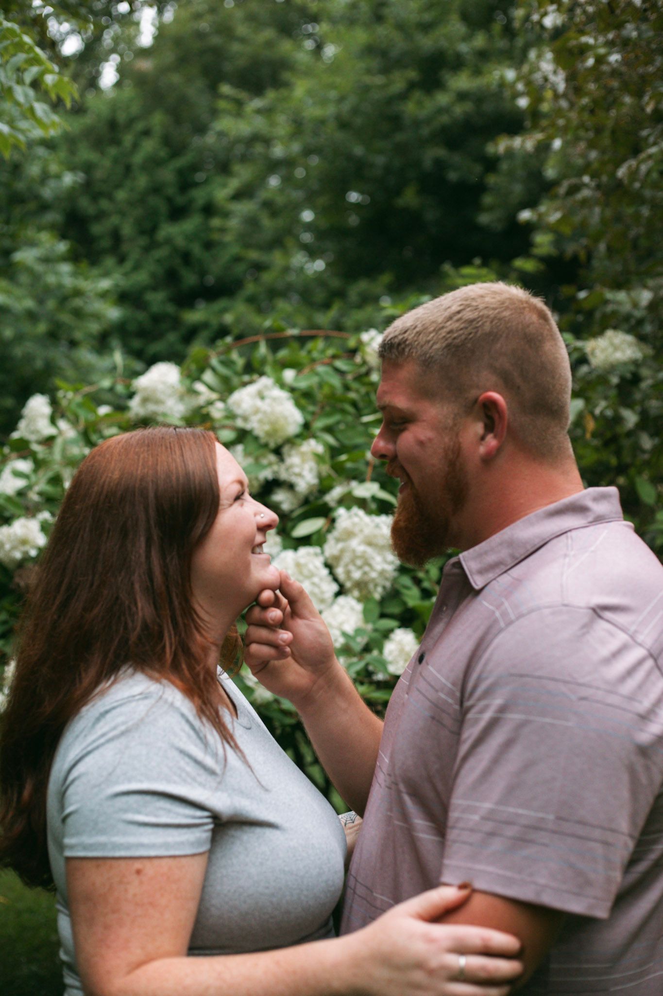 Happy engaged couple gazes at each other lovingly in front of white hydrangea blooms outdoors.