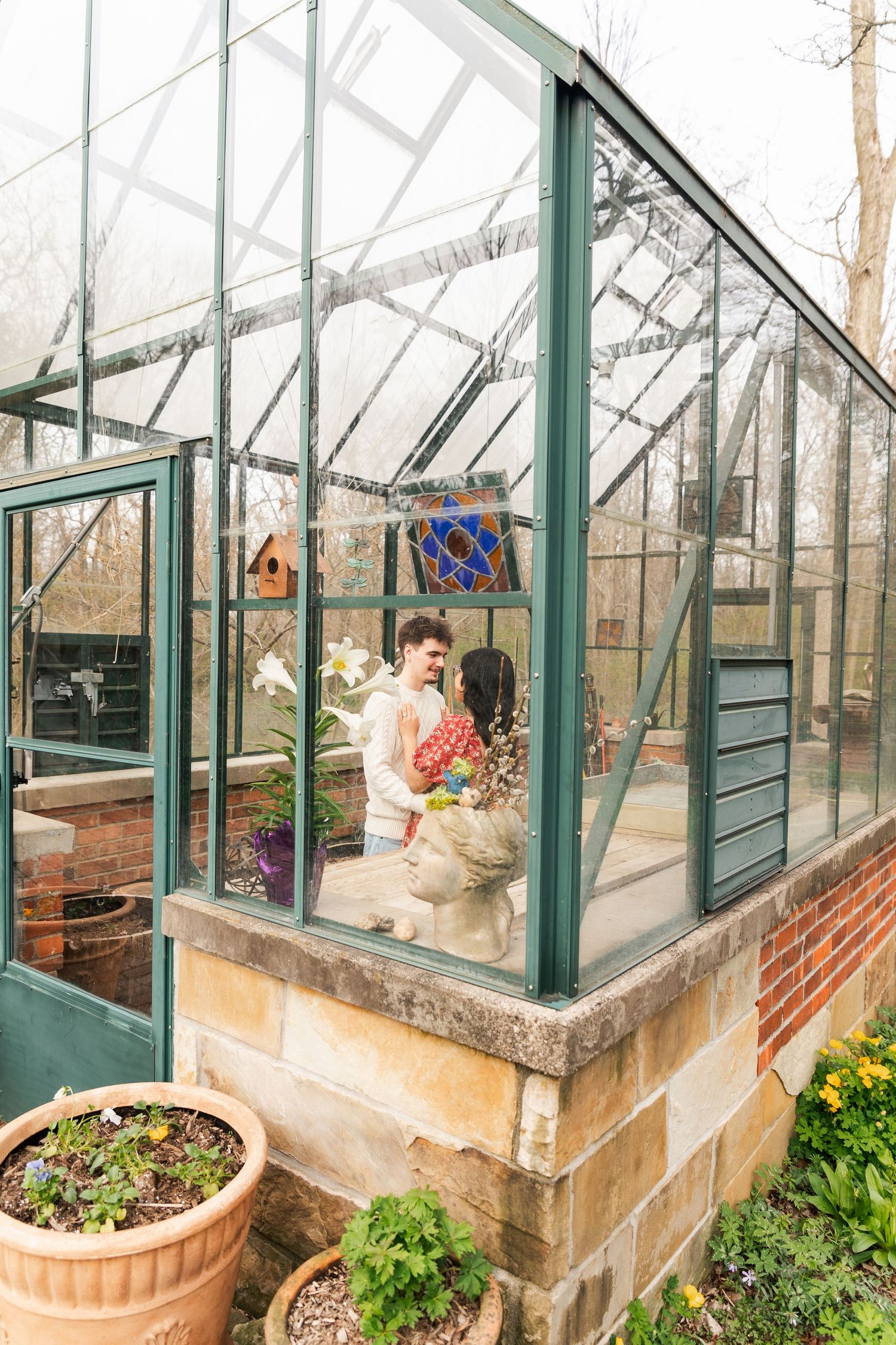 Couple shares a romantic moment inside a charming glass greenhouse surrounded by potted plants and flowers.