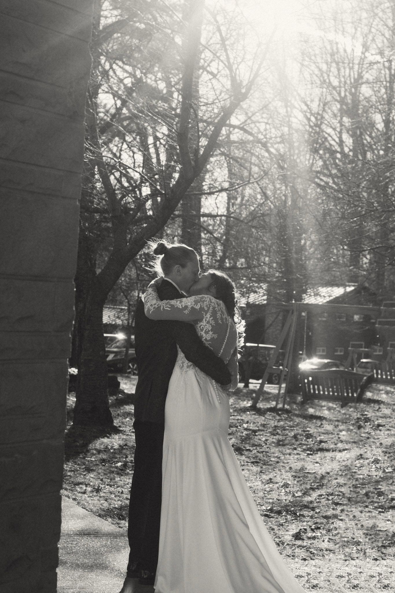 Black and white photo of a couple kissing in sunlit trees, bride in white gown.
