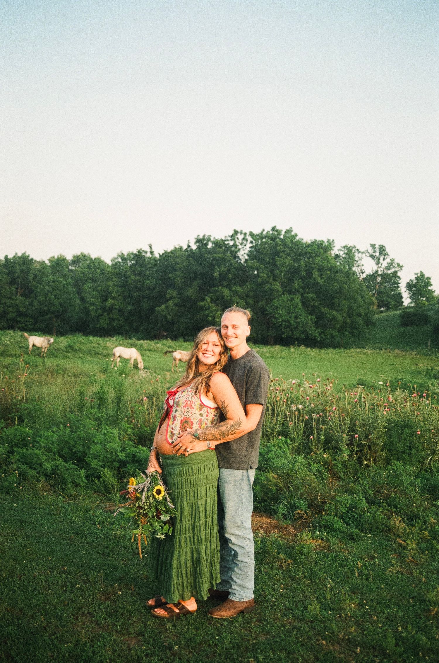 Pregnant couple smiling in a green field with cows grazing, woman holding sunflowers, wearing a green skirt at golden hour.