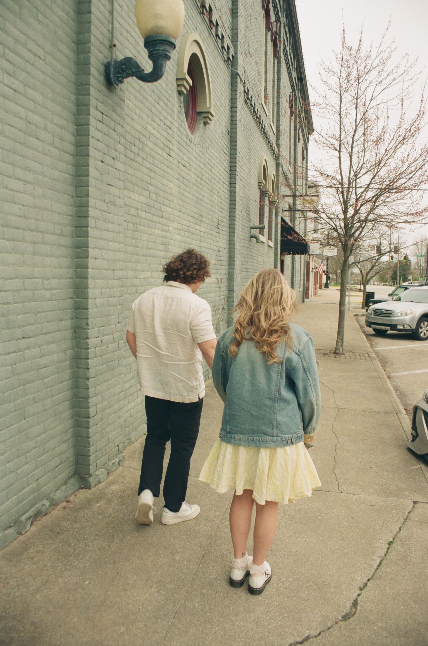 Couple walking together along a brick building sidewalk, woman in denim jacket holding man's arm.