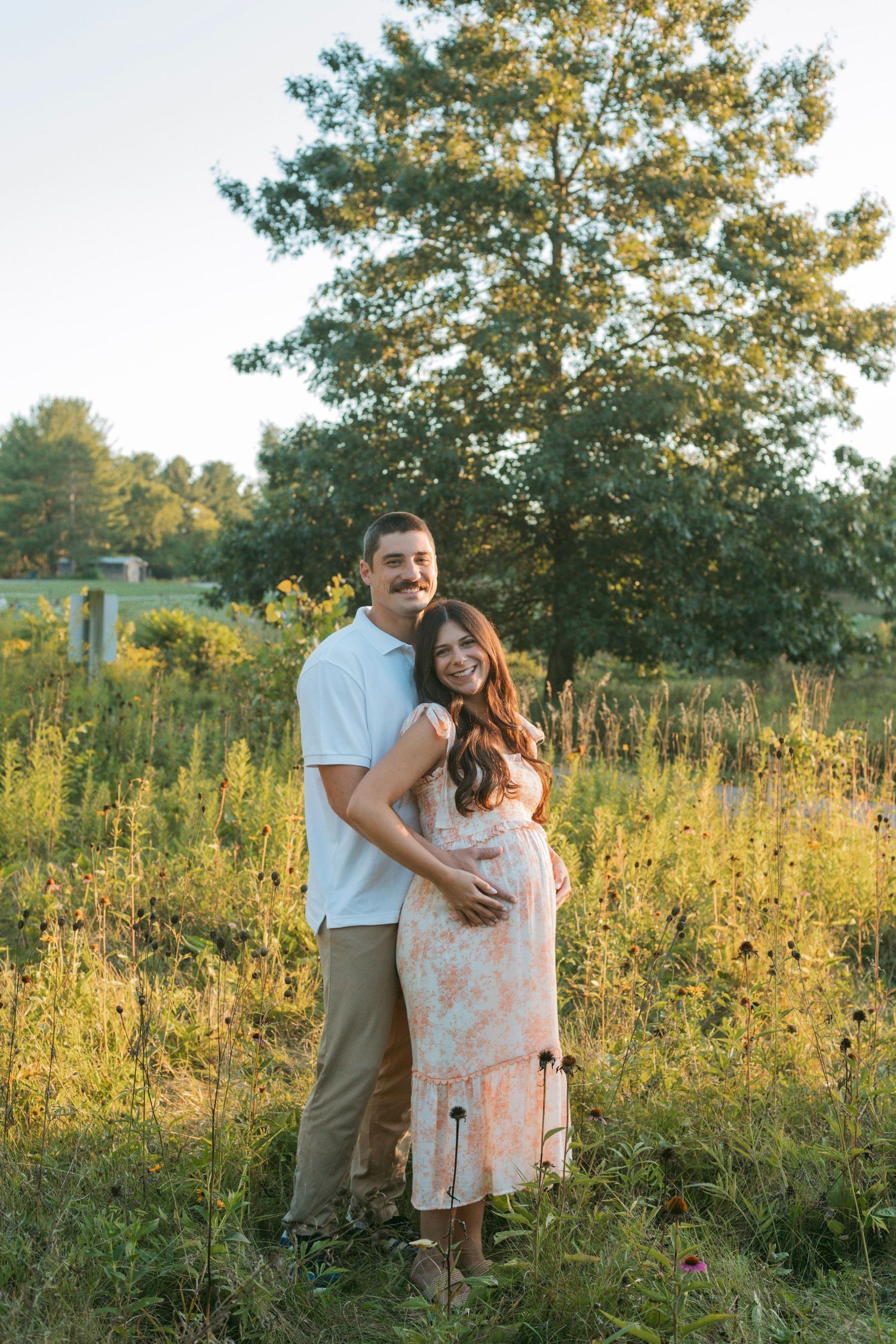 Expectant couple smiling and embracing in sunlit wildflower meadow, woman in floral maternity dress.