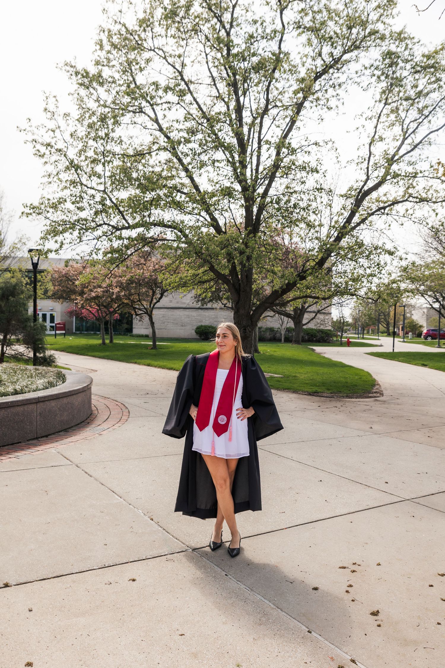 Female graduate in black graduation gown with red honor sash walking on college campus pathway.