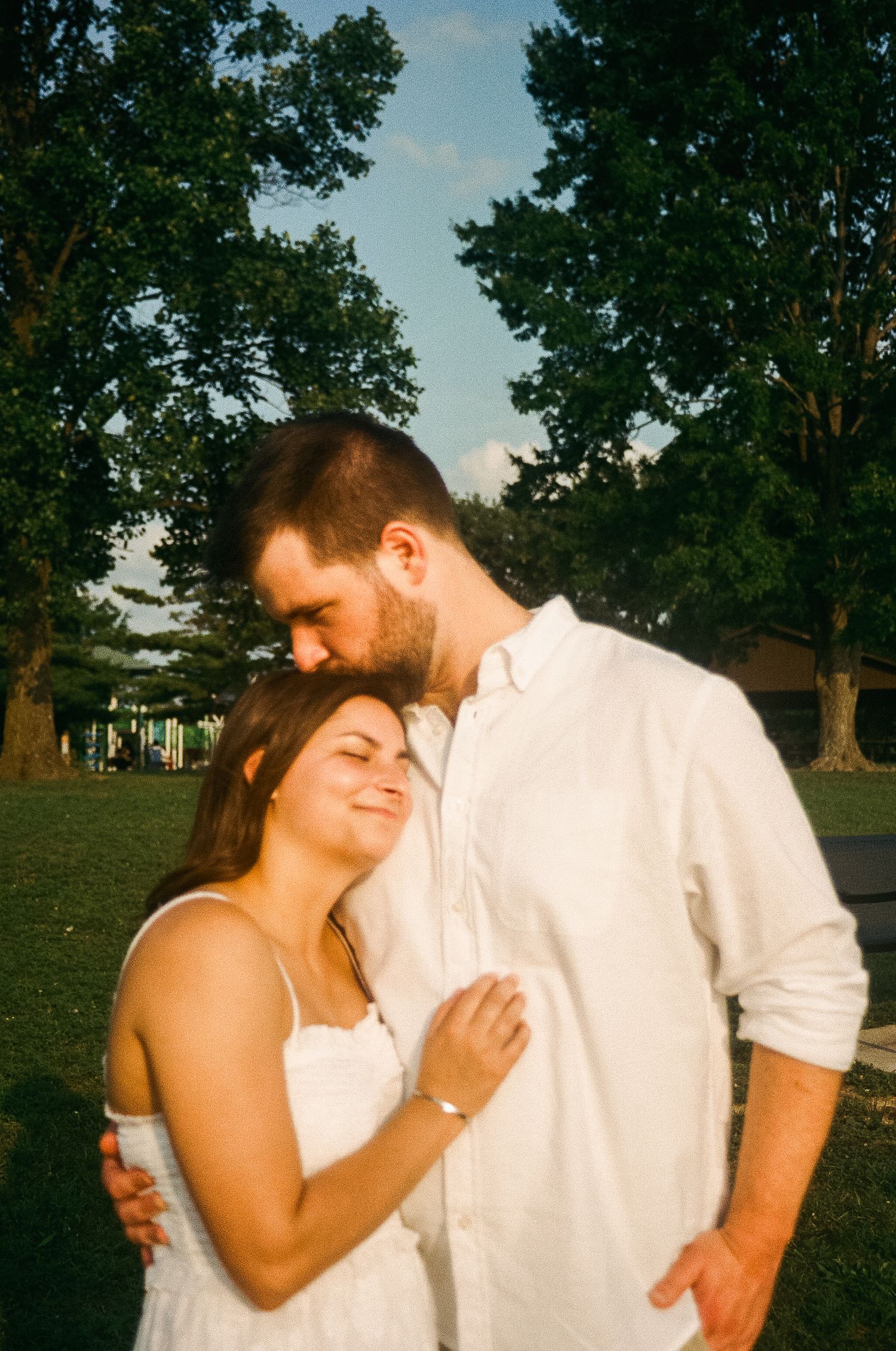 Couple in white outfits sharing a tender moment outdoors, man kissing woman's forehead near lush green trees.
