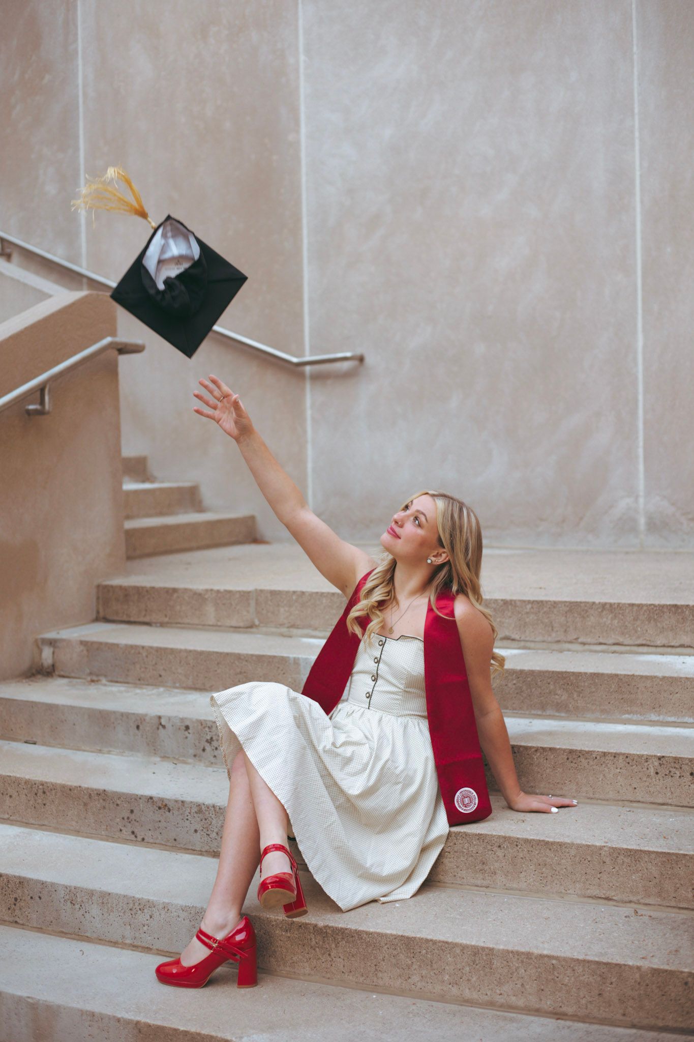 Blonde graduate in white dress tossing her cap on marble stairs wearing red heels and sash.