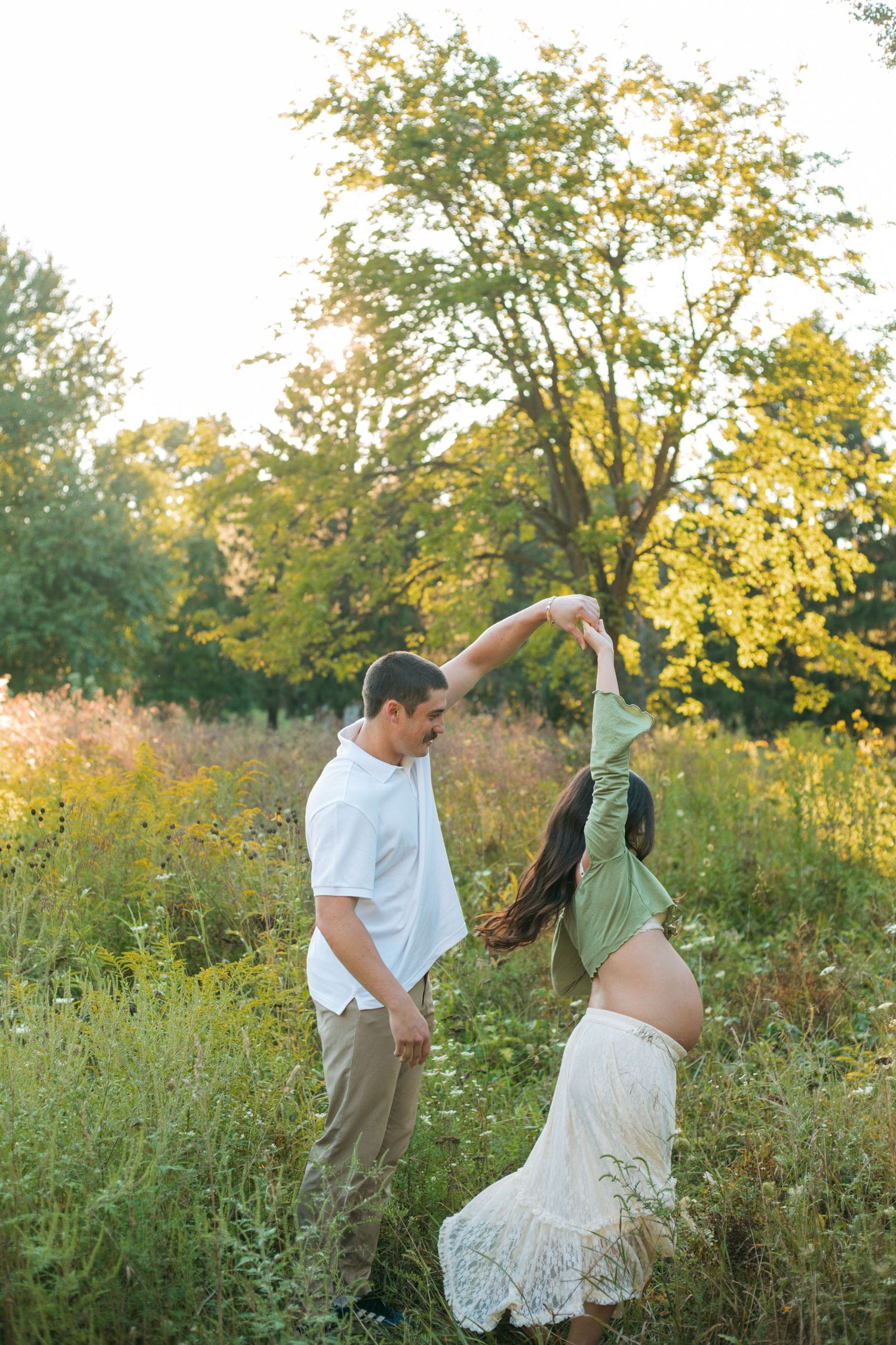 Joyful pregnant couple dancing outdoors, man spinning woman in green top in a golden sunlit field.