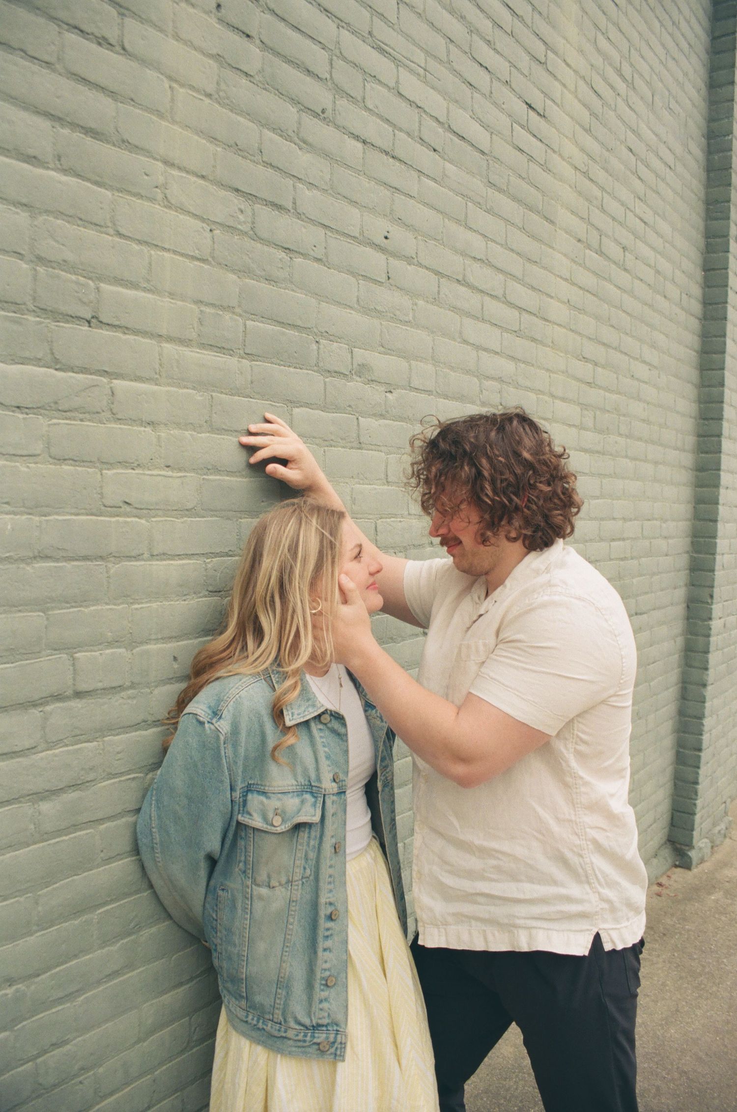 Couple in a tender moment against a green brick wall, man leaning over woman affectionately.