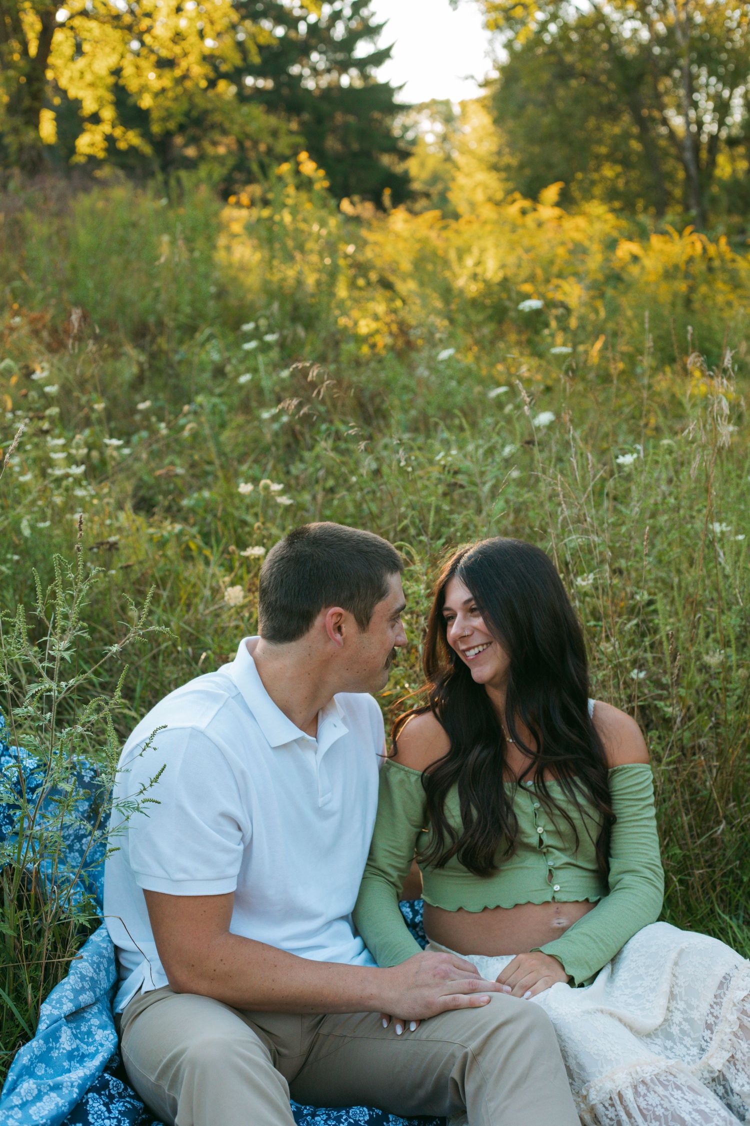 Expecting couple sitting together on blanket in golden meadow, smiling and holding hands.