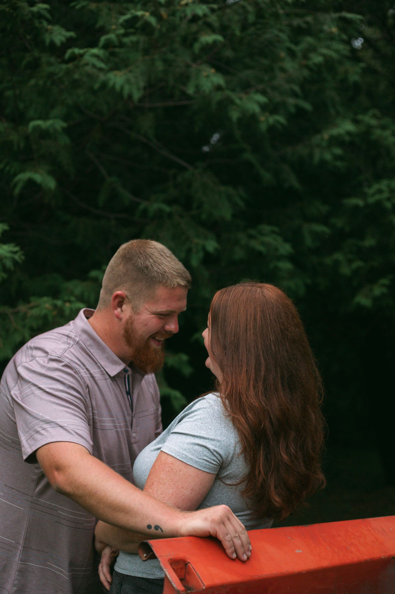 Smiling couple shares a tender moment leaning against red railing surrounded by lush green trees.
