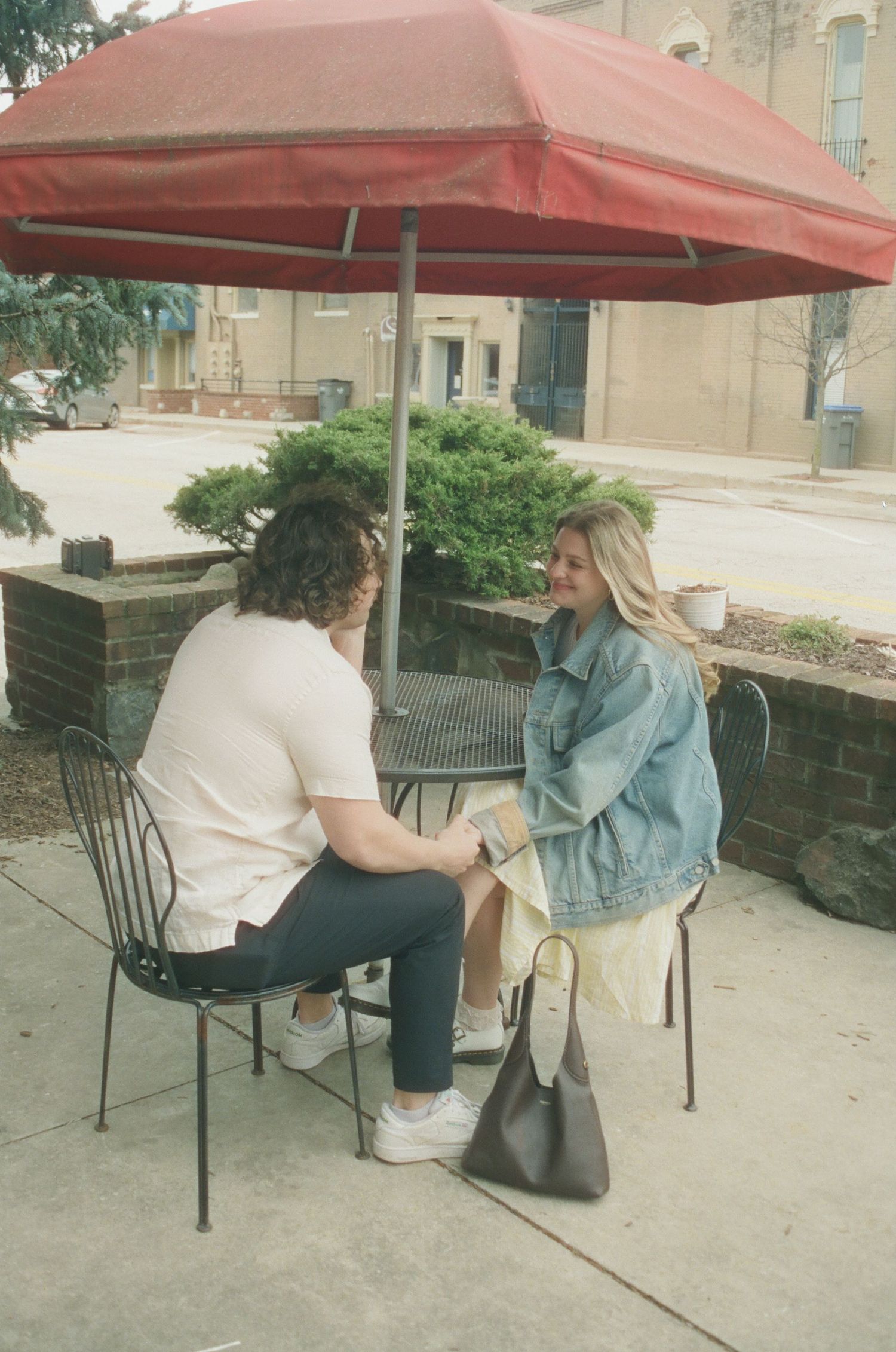 Couple sitting together under red patio umbrella at outdoor cafe, holding hands and smiling.