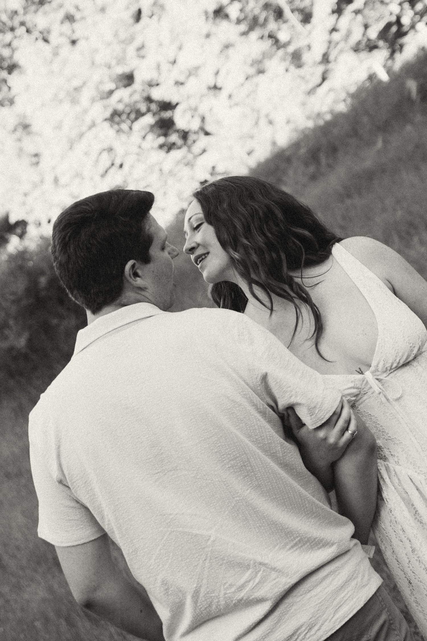 Black and white photo of couple embracing and laughing together outdoors under bright sunlit tree canopy.