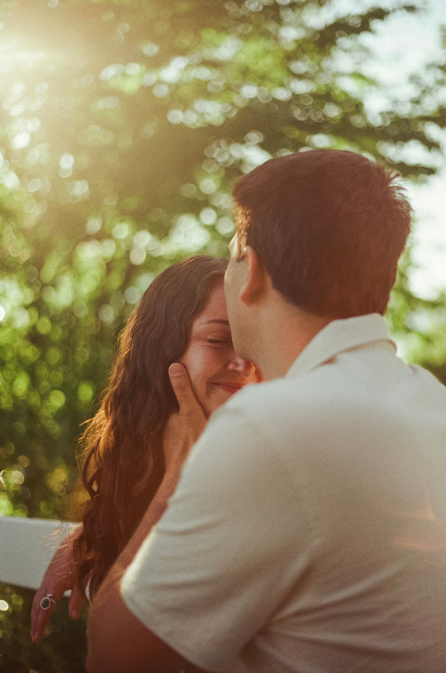 A couple shares a tender kiss bathed in golden sunlight with lush green trees in the background.