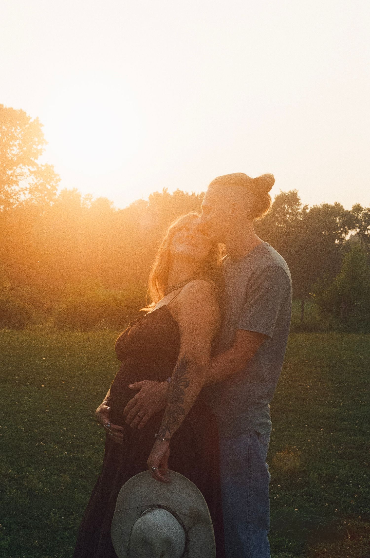 Couple embracing at golden hour sunset in a field, man kissing woman's cheek as she smiles, maternity photo.