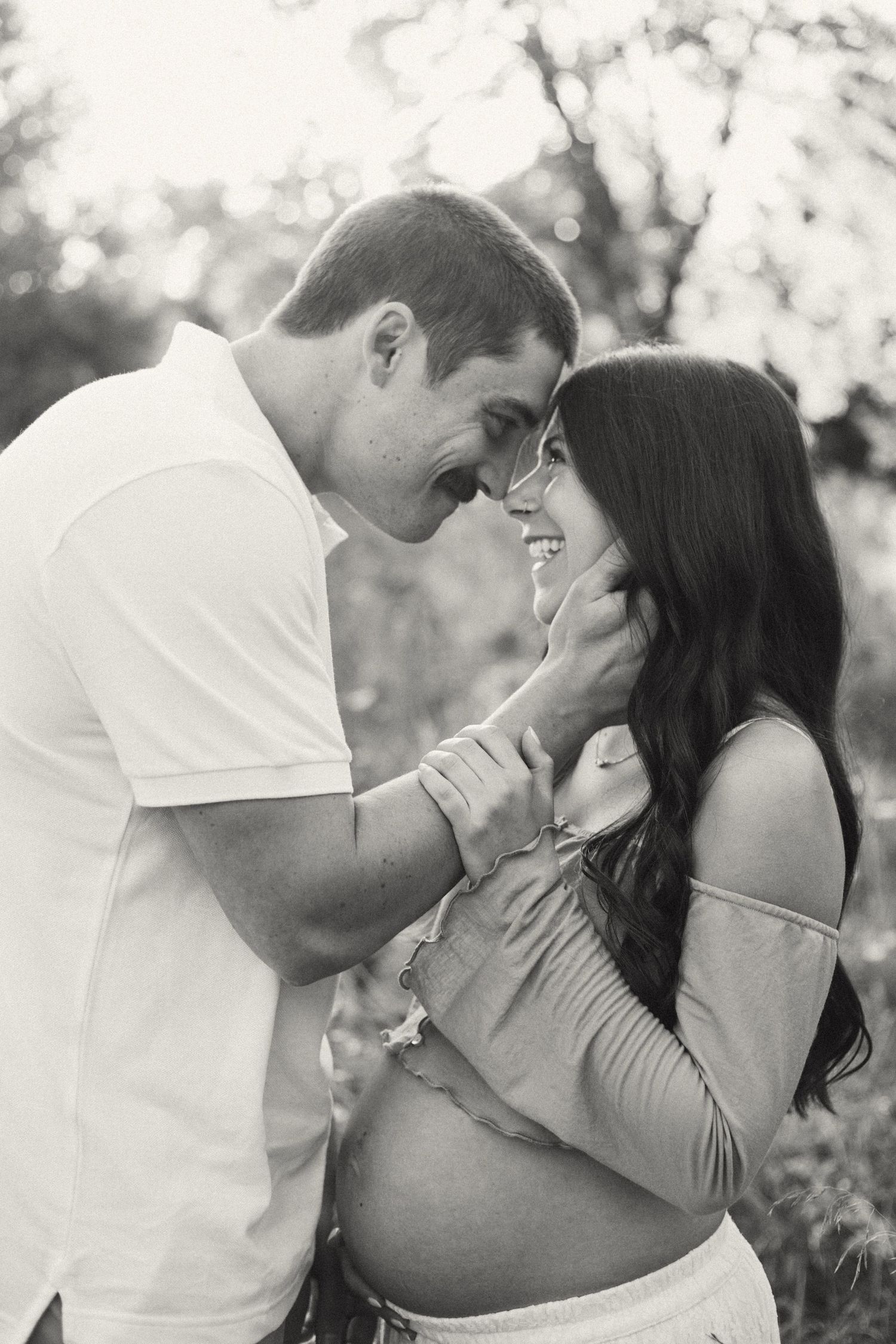 Black and white photo of expectant couple laughing together in a field, foreheads nearly touching.