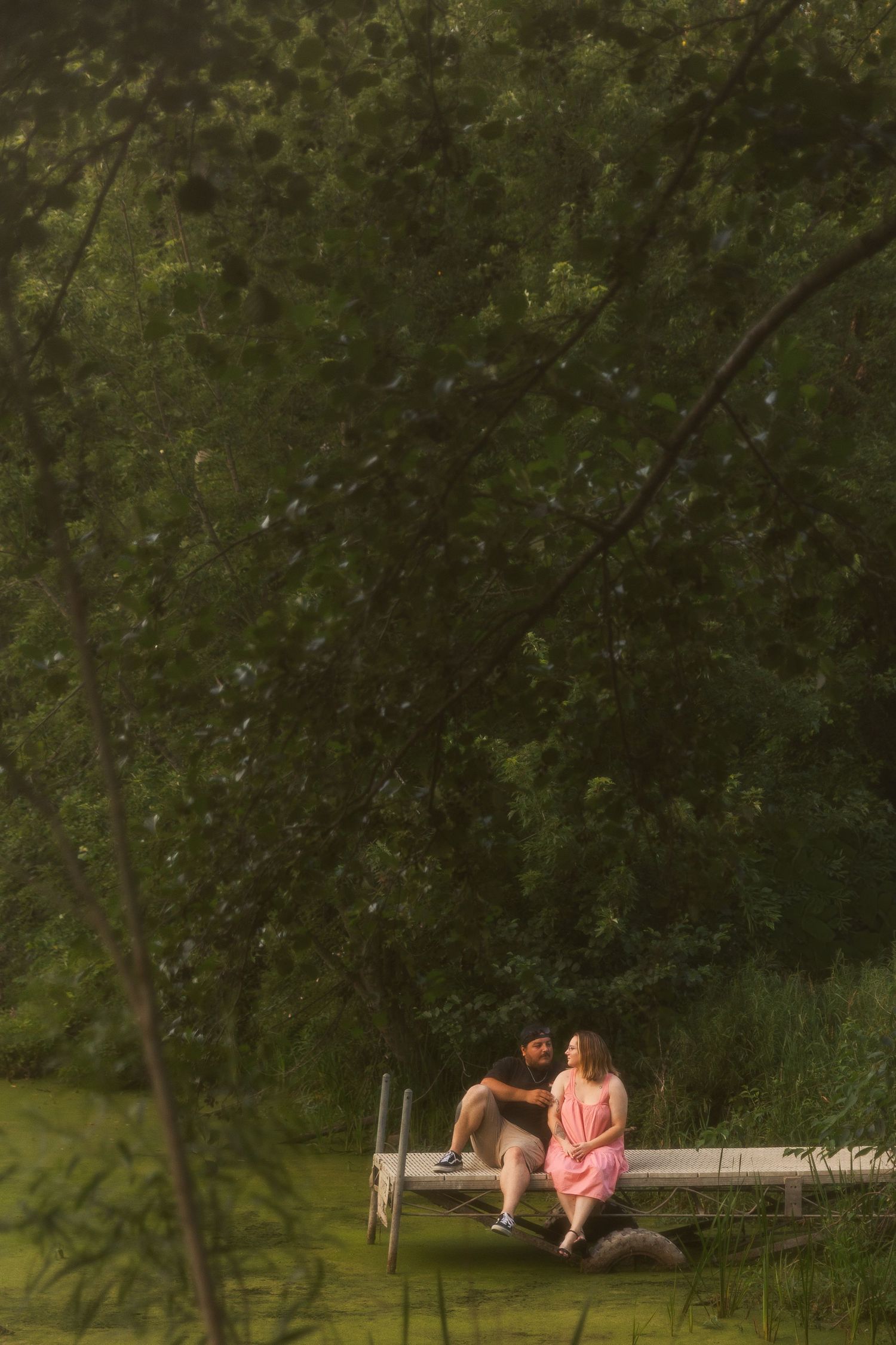 Couple sitting together on a wooden dock surrounded by lush green trees in a romantic outdoor setting.