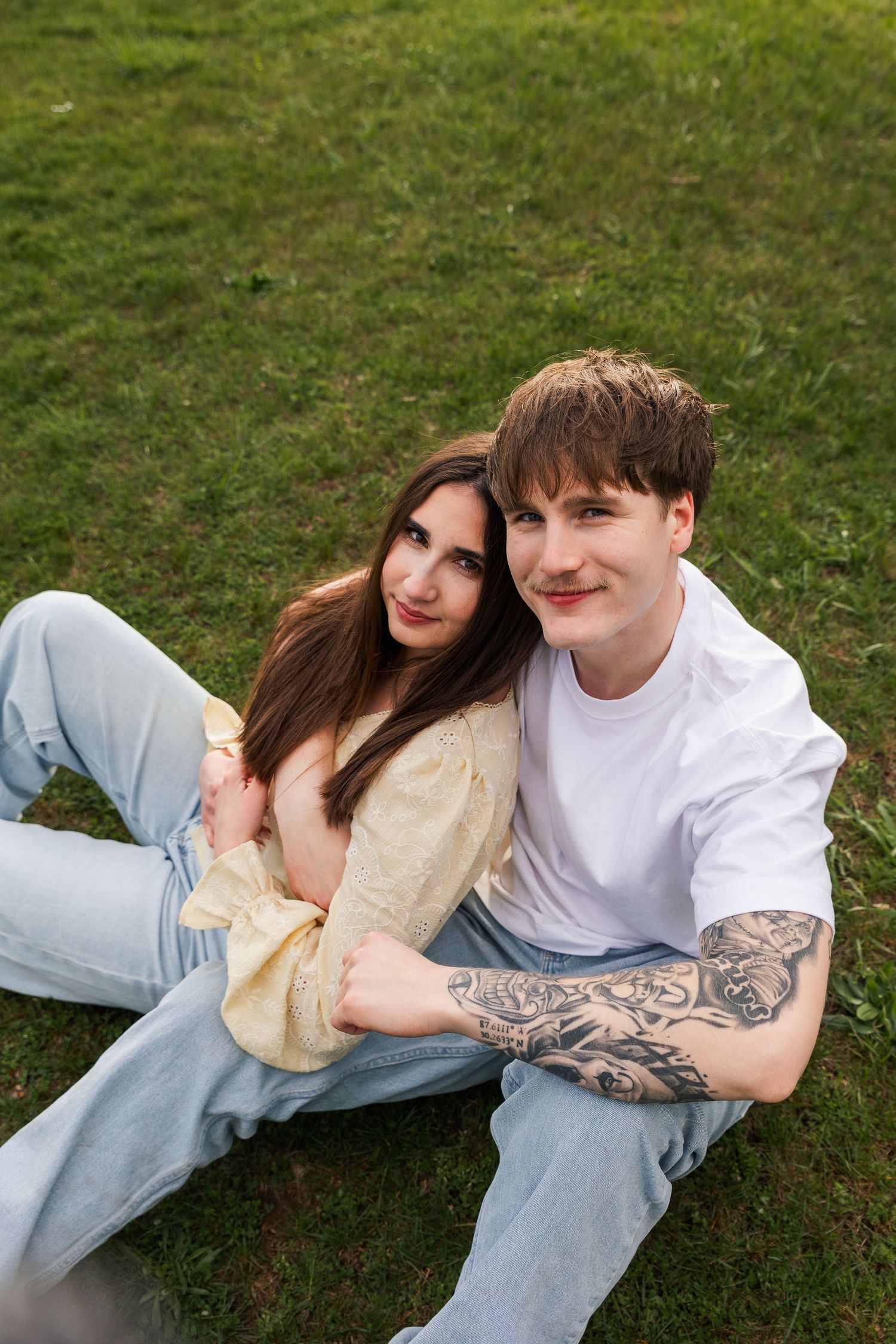 Young couple sitting together on green grass, man with tattoos in white tee embracing woman.