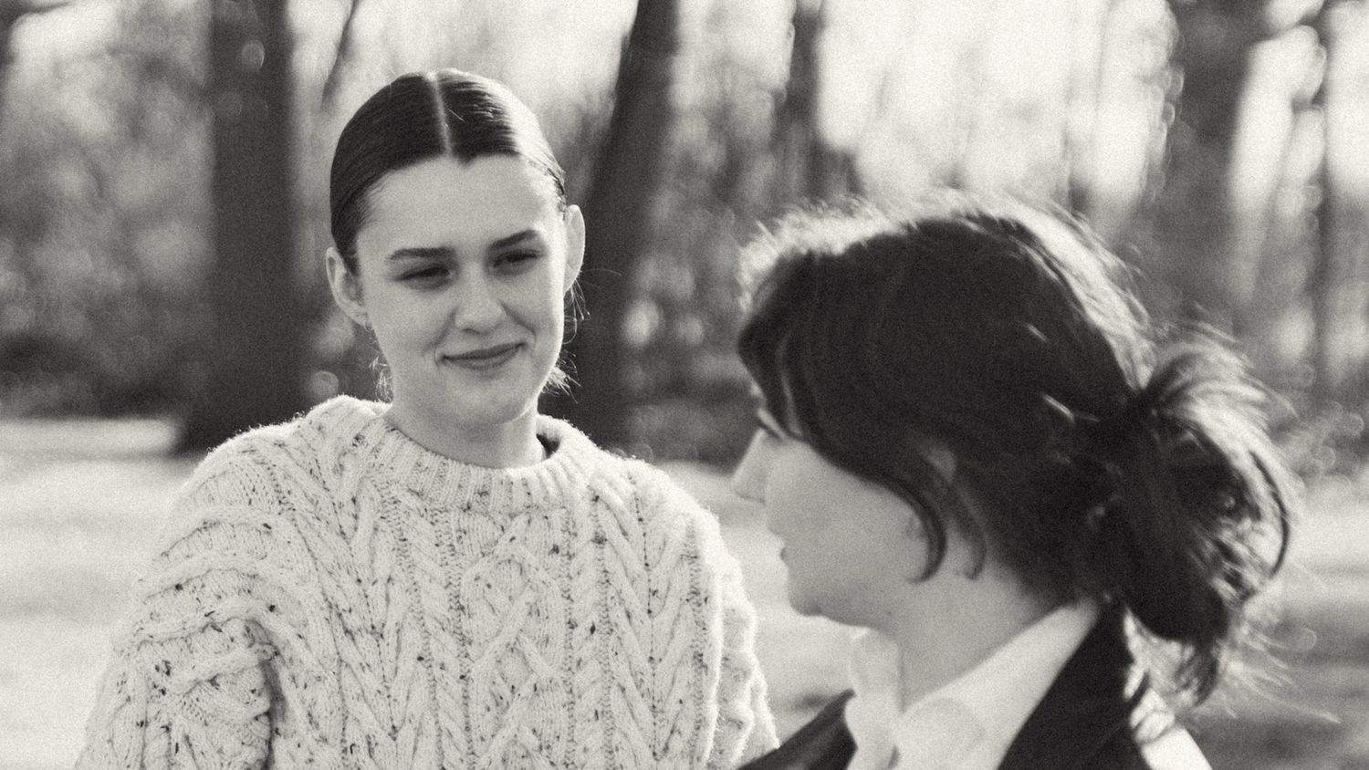 A black and white portrait of two young women smiling softly at each other outdoors in a wooded setting.