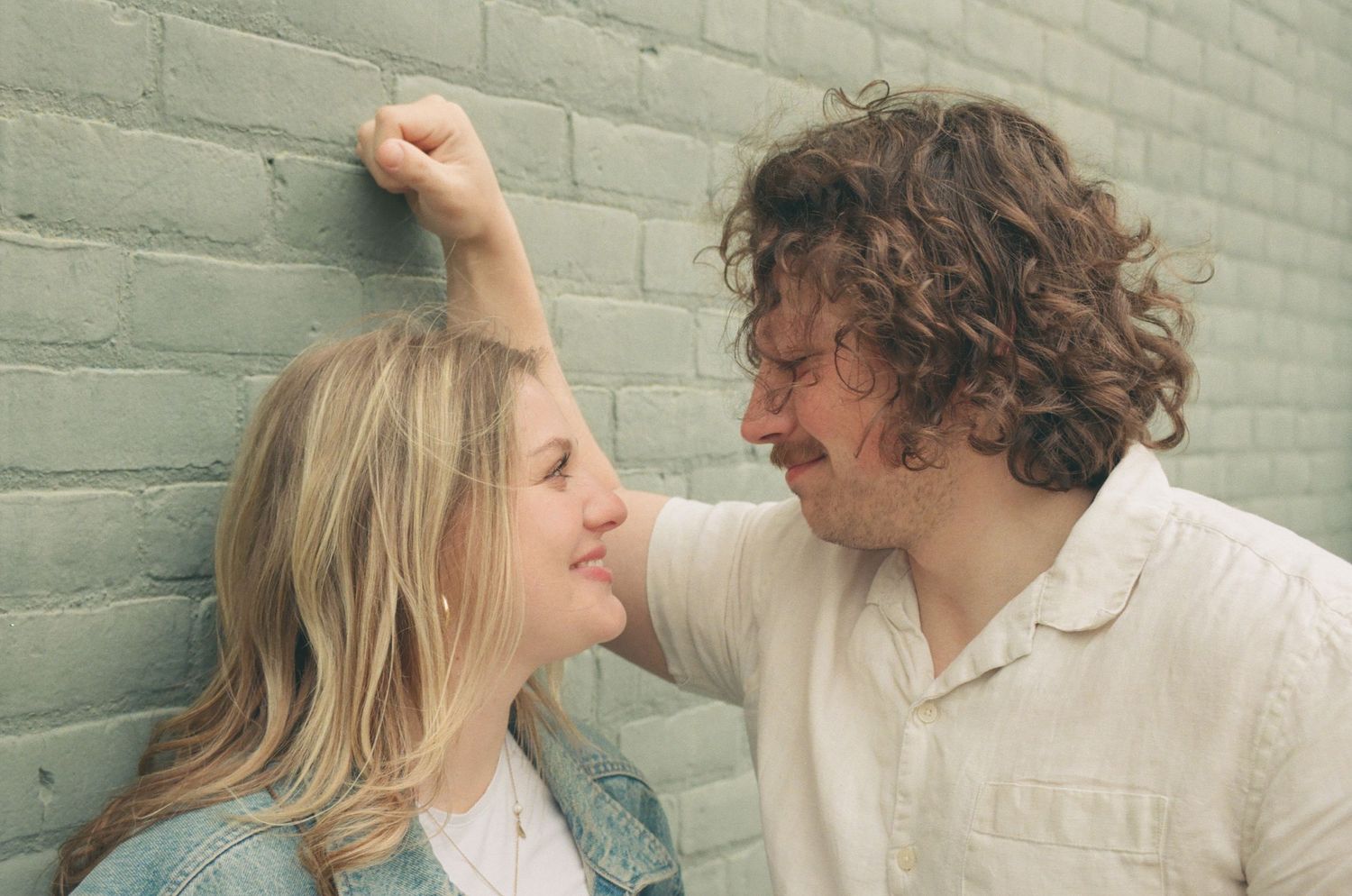 Playful couple laughing against a green brick wall, man holding his fist above womans head.