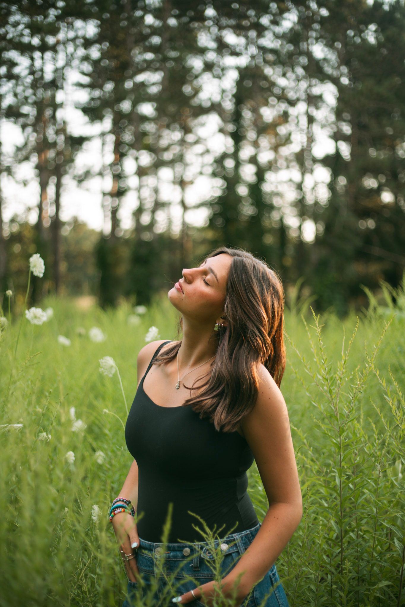 Young woman in black tank top standing in tall wildflower meadow with pine forest backdrop.