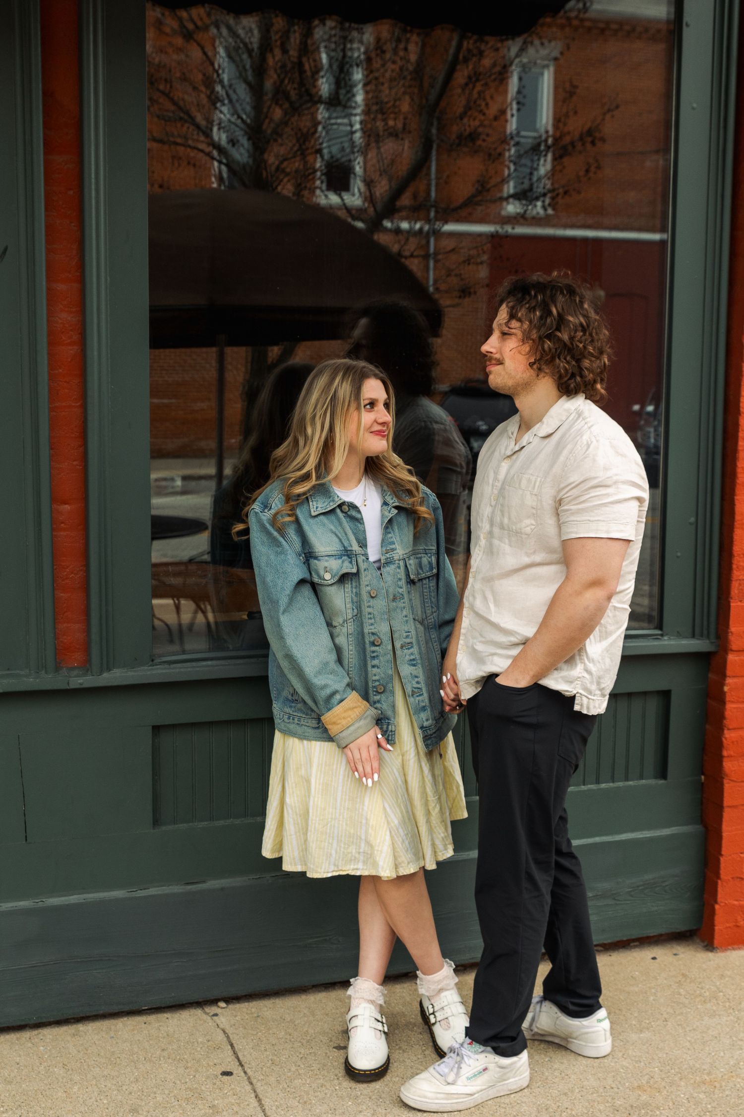 Couple holds hands and gazes at each other standing in front of a green storefront on a sidewalk.