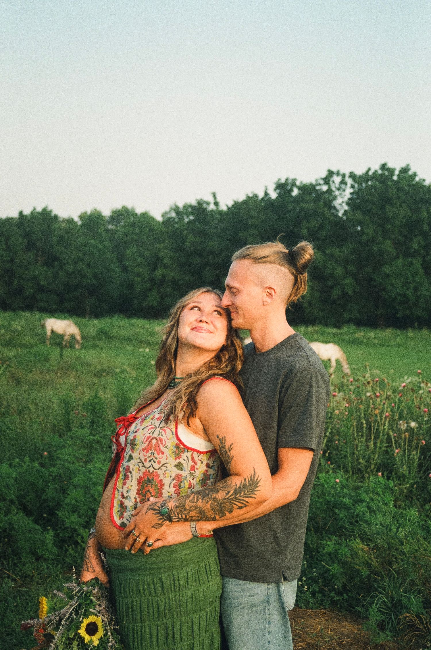 Couple embracing in a green field at golden hour, woman with tattoos holding sunflowers, horse grazing in background.