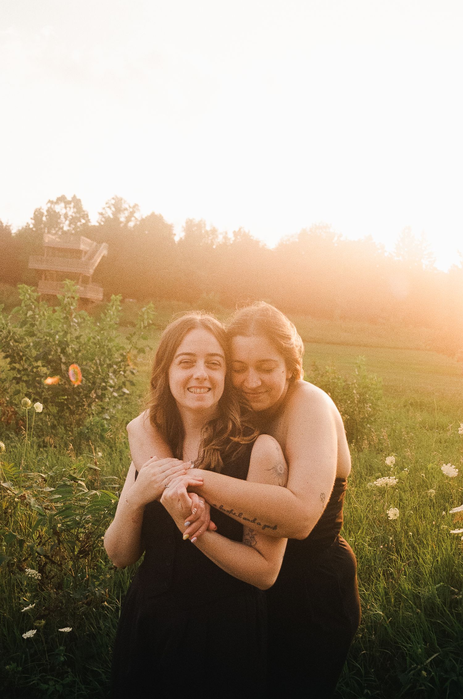 Two young women embracing and smiling in a sunlit meadow with wildflowers during golden hour.