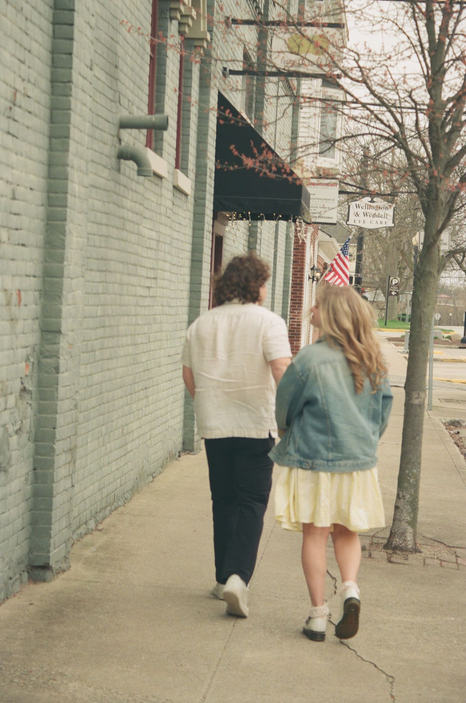 Couple walking hand in hand down a charming brick sidewalk, woman in denim jacket and yellow skirt.