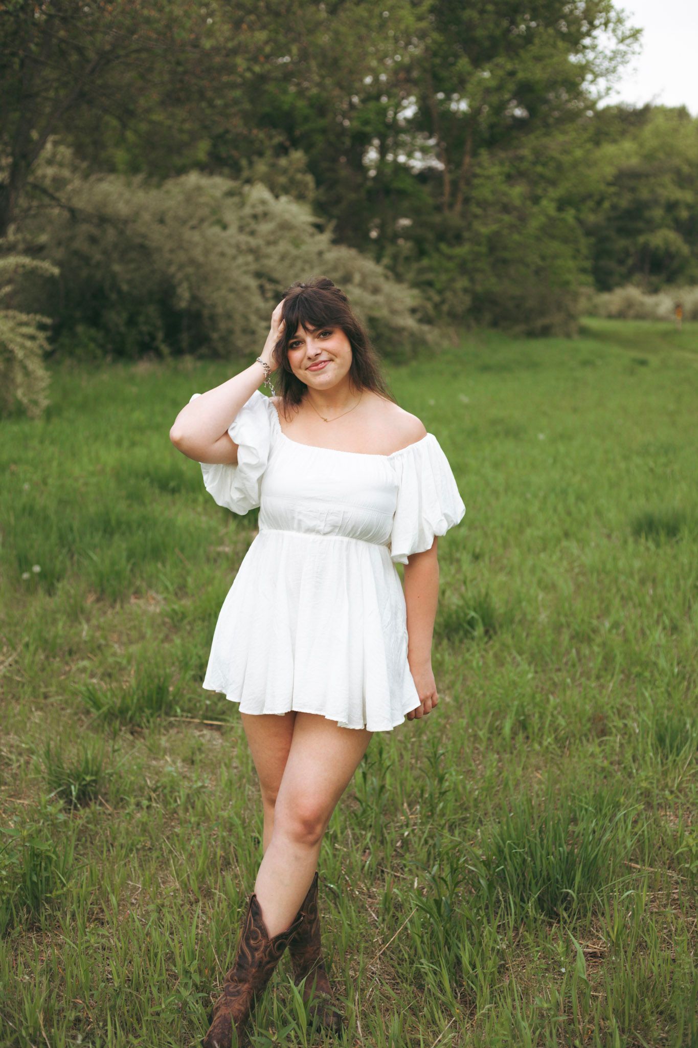 Young woman in white off-shoulder mini dress and brown cowboy boots in green field.