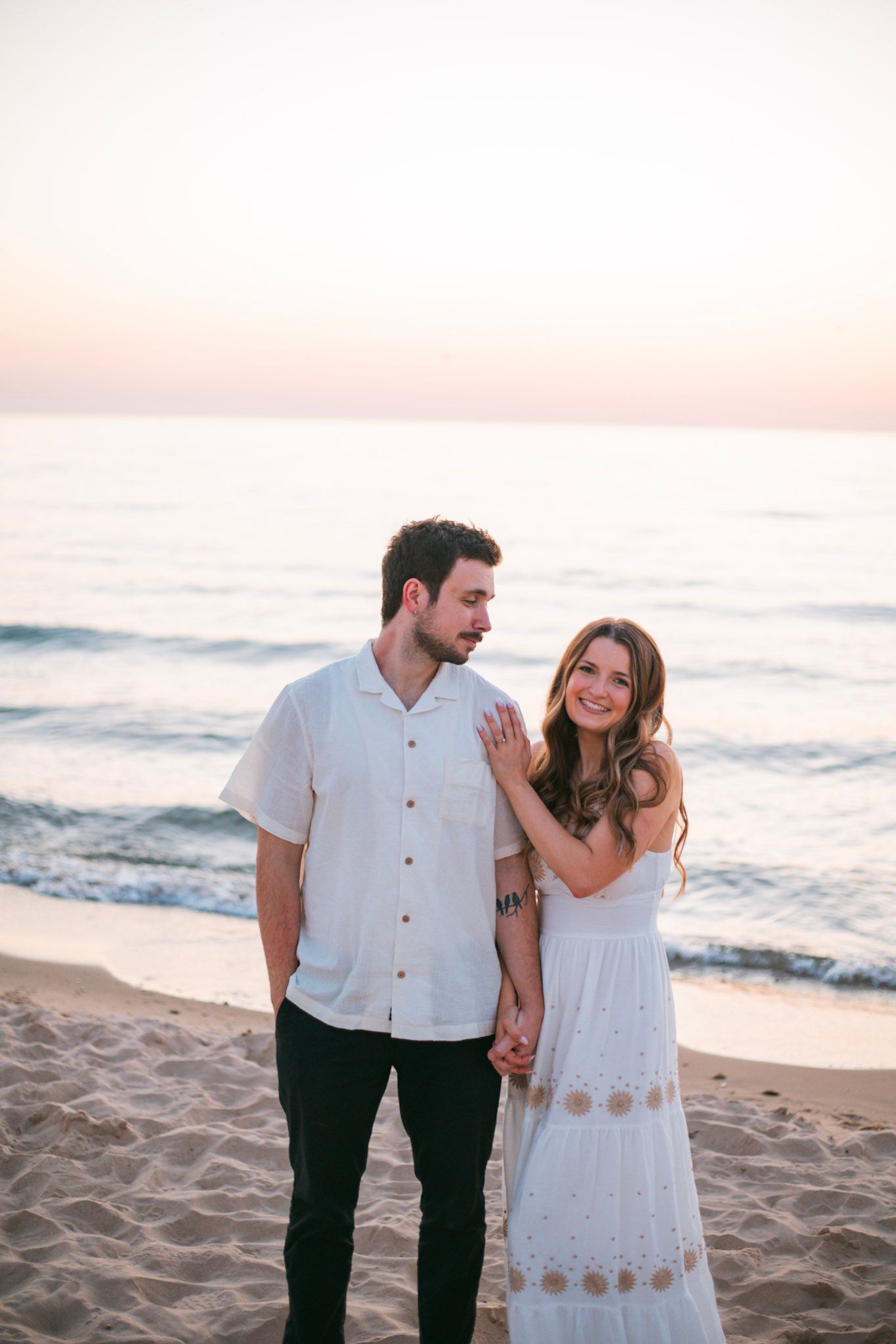 Romantic couple holds hands on sandy beach at sunset, woman in white dress smiling warmly.