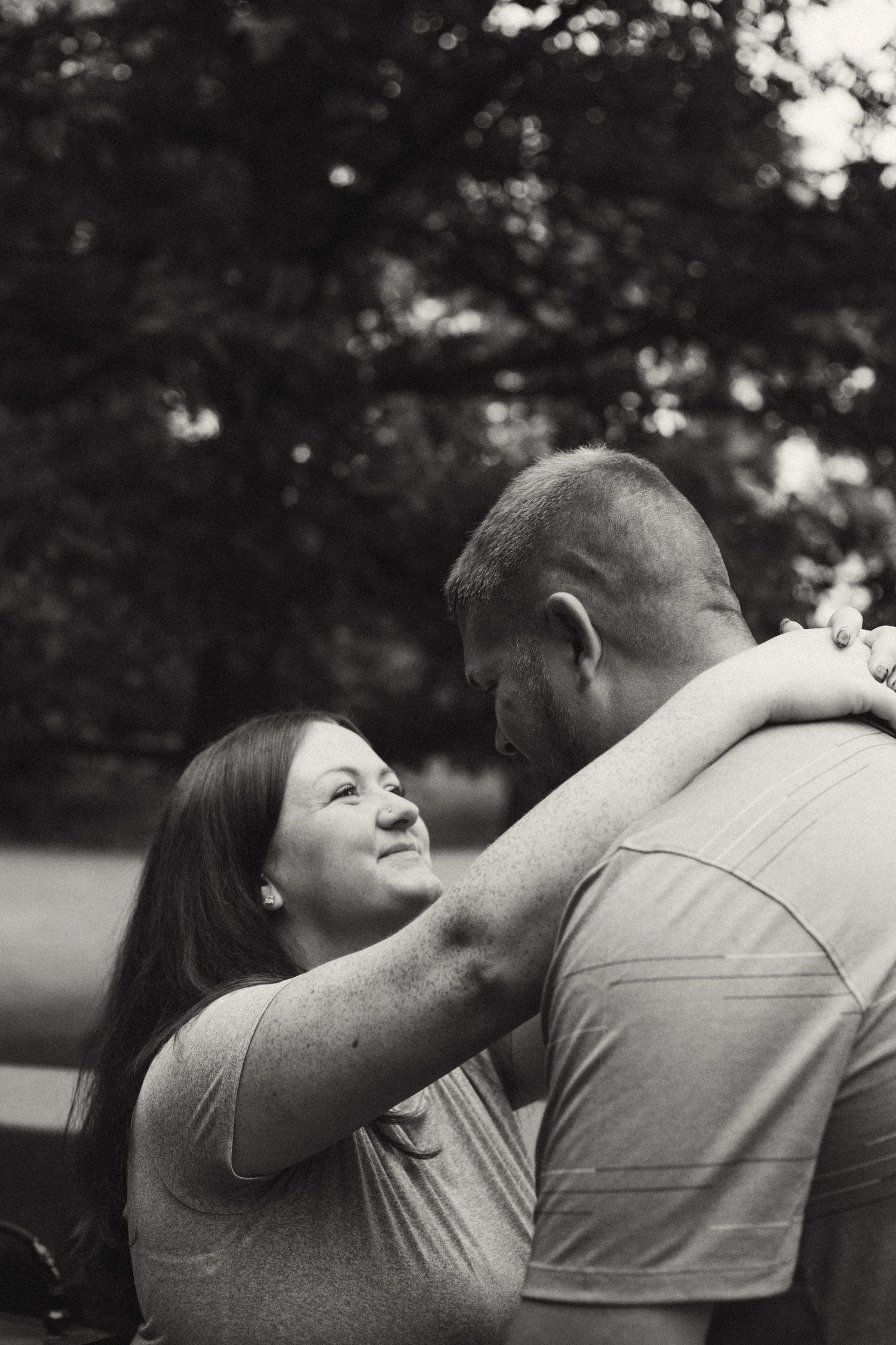 A black and white photo of a smiling woman embracing a man outdoors with softly blurred trees in the background.
