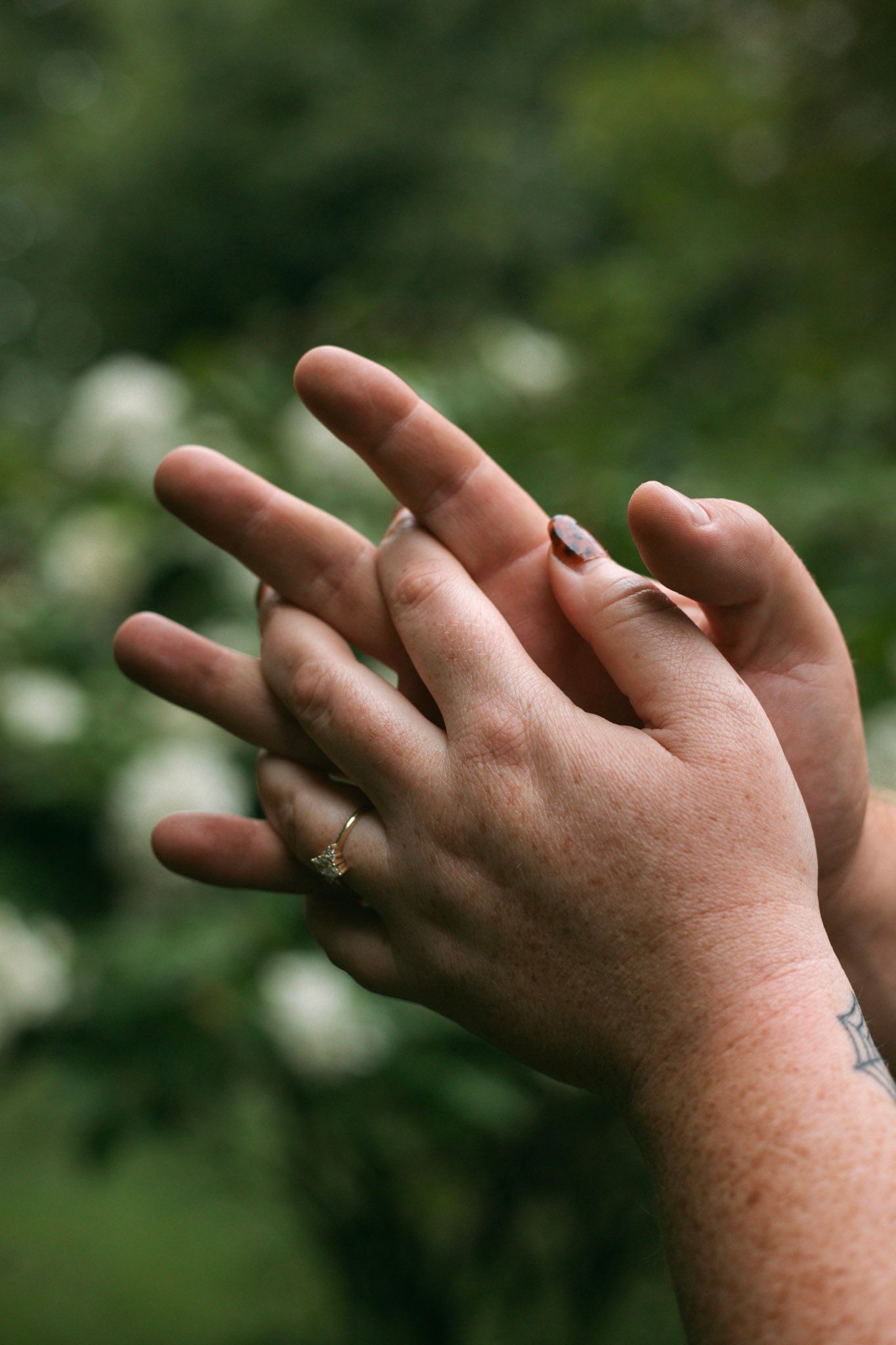 Close-up of two hands touching showing engagement ring among white flowering garden backdrop.