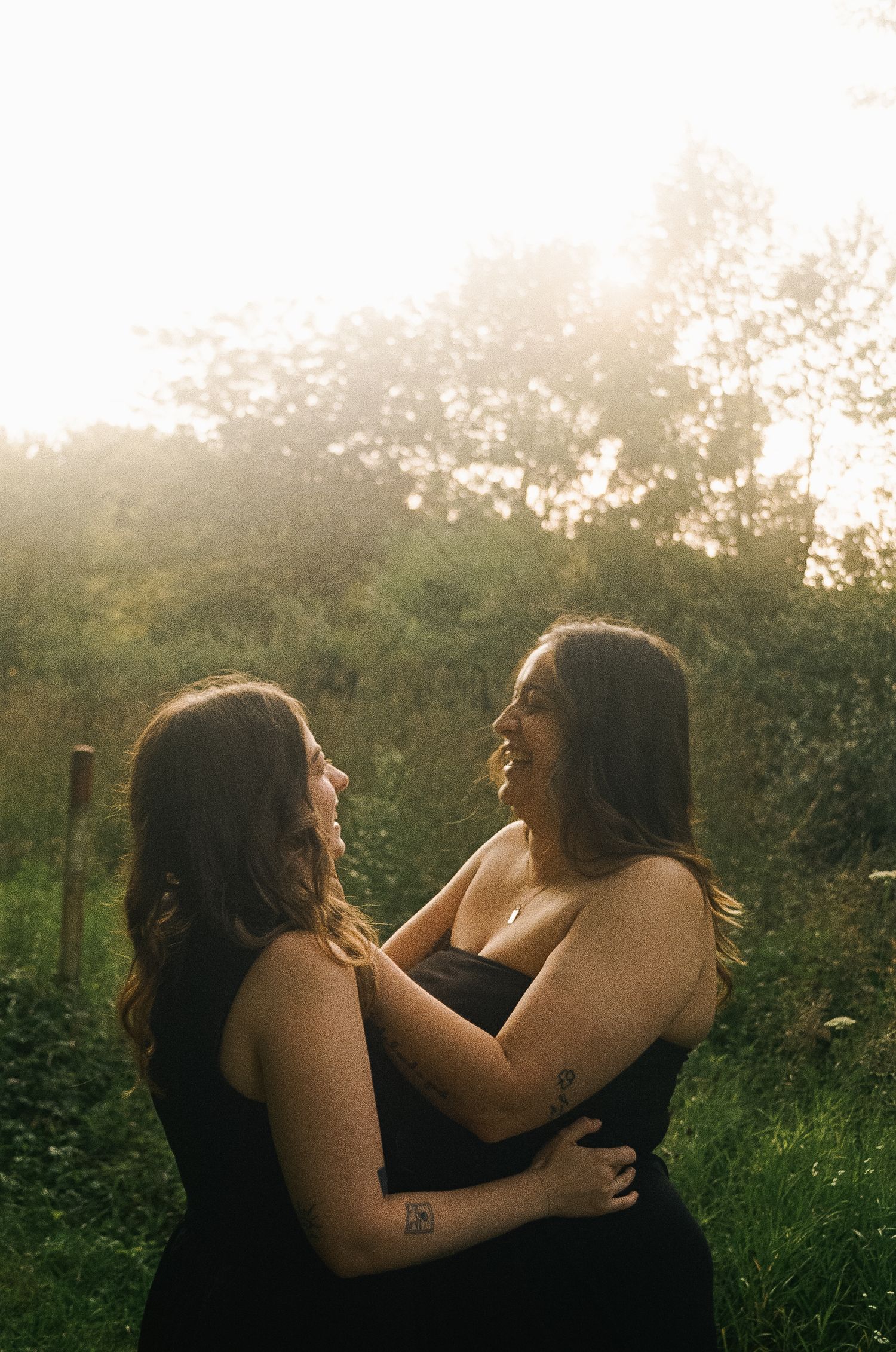 Two women in black dresses embrace and smile at each other in a sunlit garden setting.