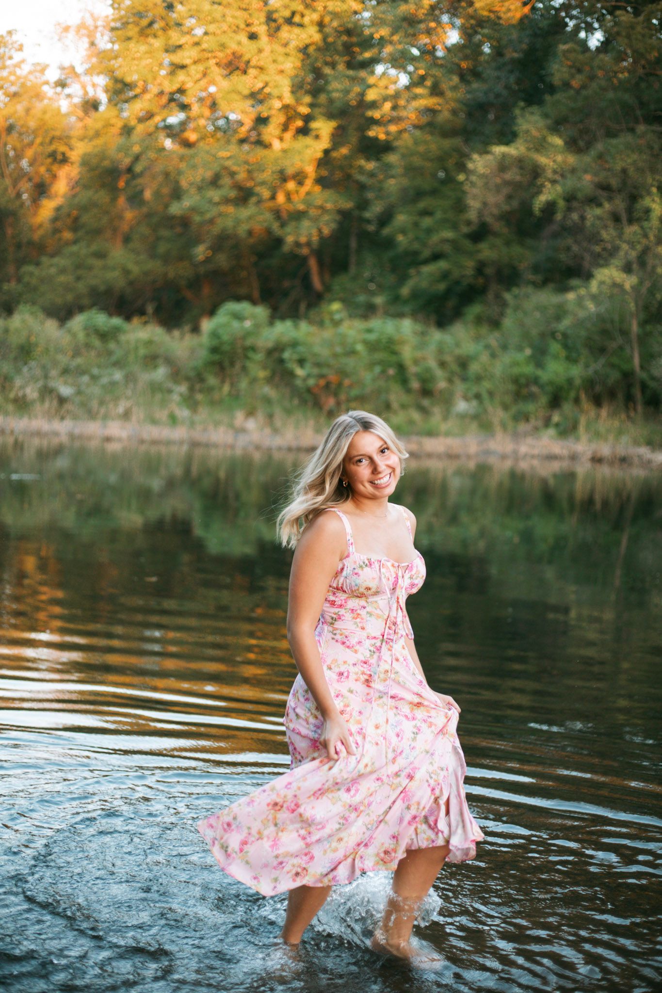 Smiling blonde woman in floral pink dress wading through a calm river surrounded by autumn trees.