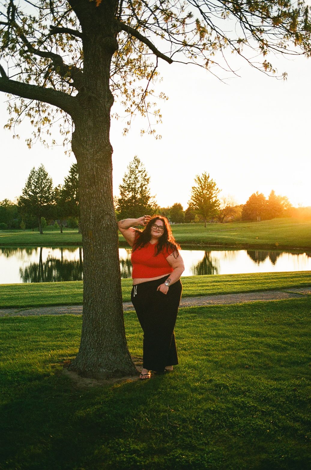 Woman in red top and black pants poses by a tree at golden hour near a serene pond.