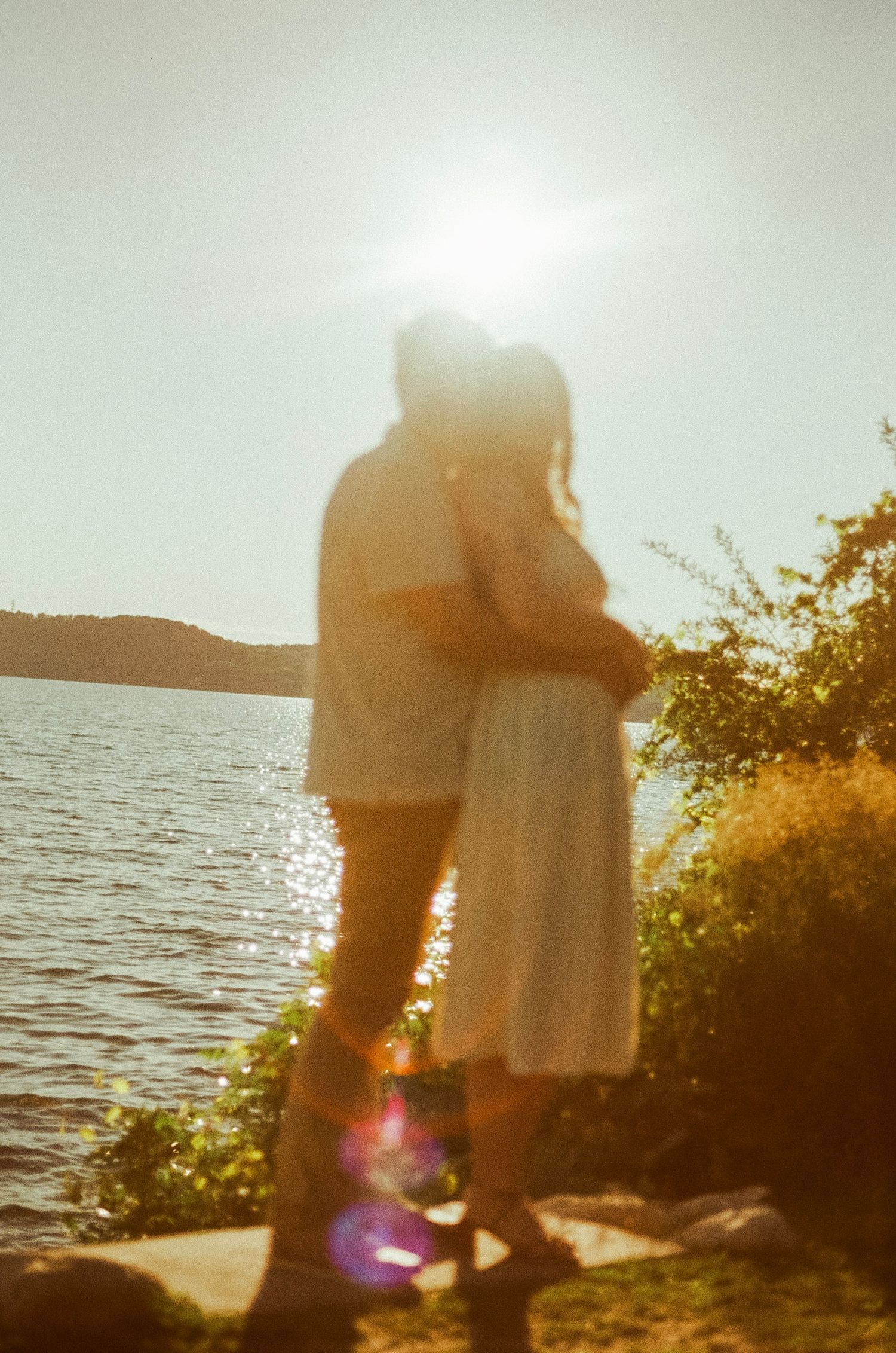 Couple embracing by a sunlit lake at golden hour with lens flare and lush greenery in the background.