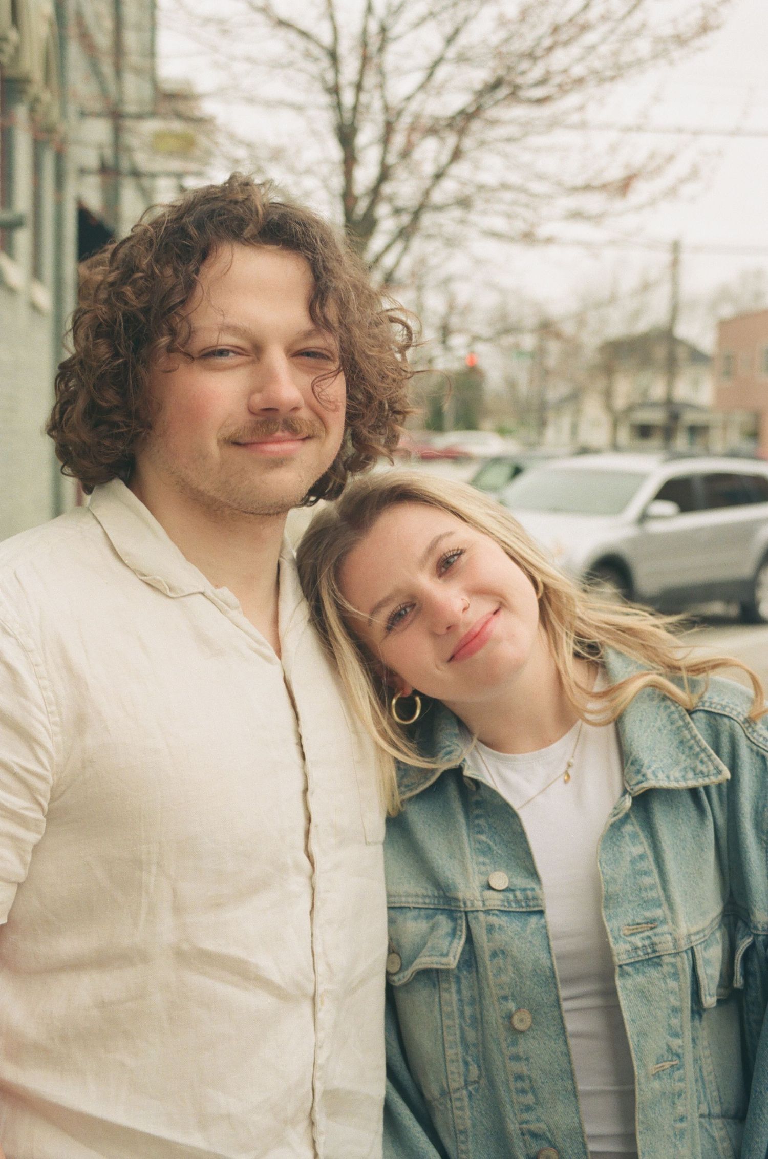 Smiling couple posing together outdoors, man with curly hair in linen shirt, woman in denim jacket.