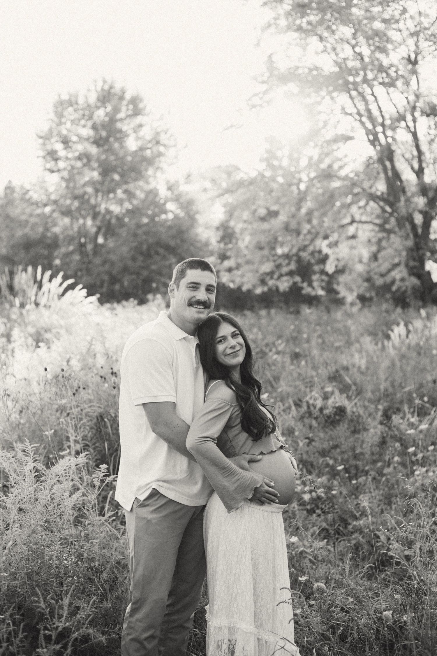 Black and white maternity photo of couple embracing in a wildflower field, man holding pregnant woman's belly.