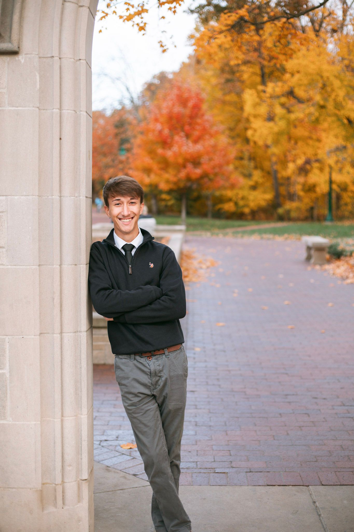 Smiling young man in black sweater and tie leaning against stone pillar with colorful autumn foliage background.
