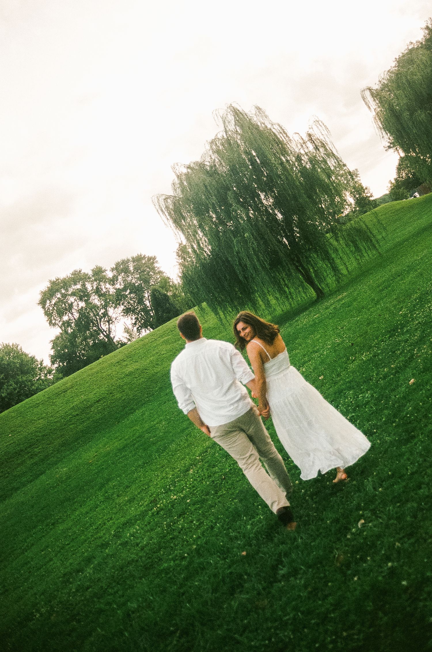 Couple in white attire holding hands while walking across lush green park with weeping willow trees.